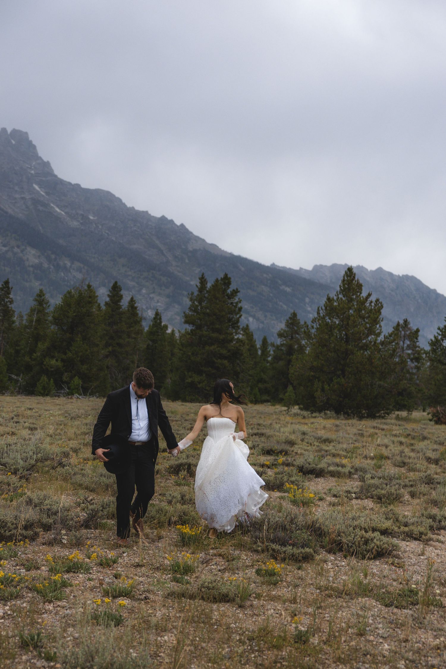 Bride and groom walking hand in hand through a mountain meadow with evergreen trees and misty peaks in the background.