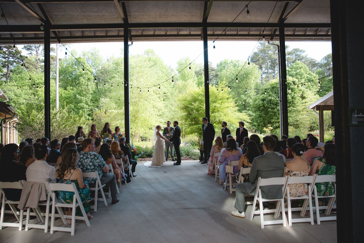 Outdoor wedding ceremony under a pavilion with white chairs and lush green trees in the background.