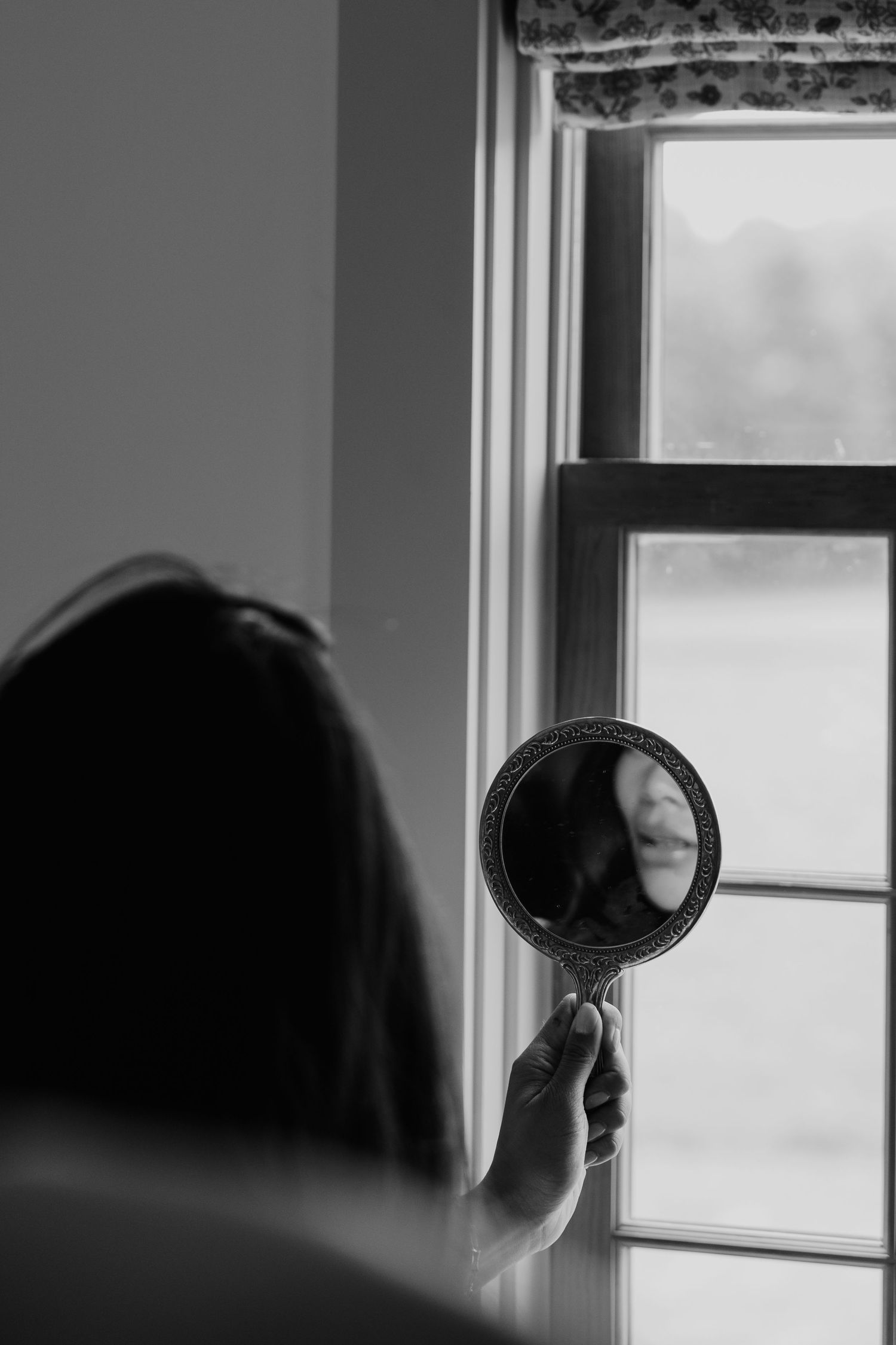 Black and white photo of a woman holding an ornate hand mirror reflecting her face near a bright window.