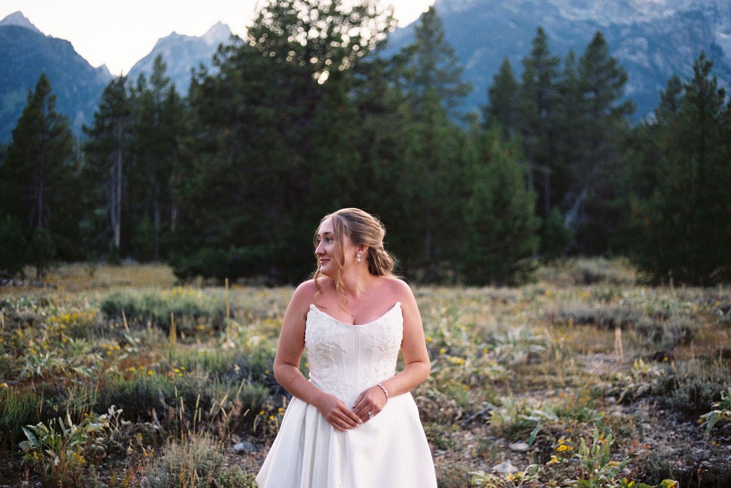 Bride in white wedding gown smiling in mountain meadow with pine trees and rocky peaks in background.