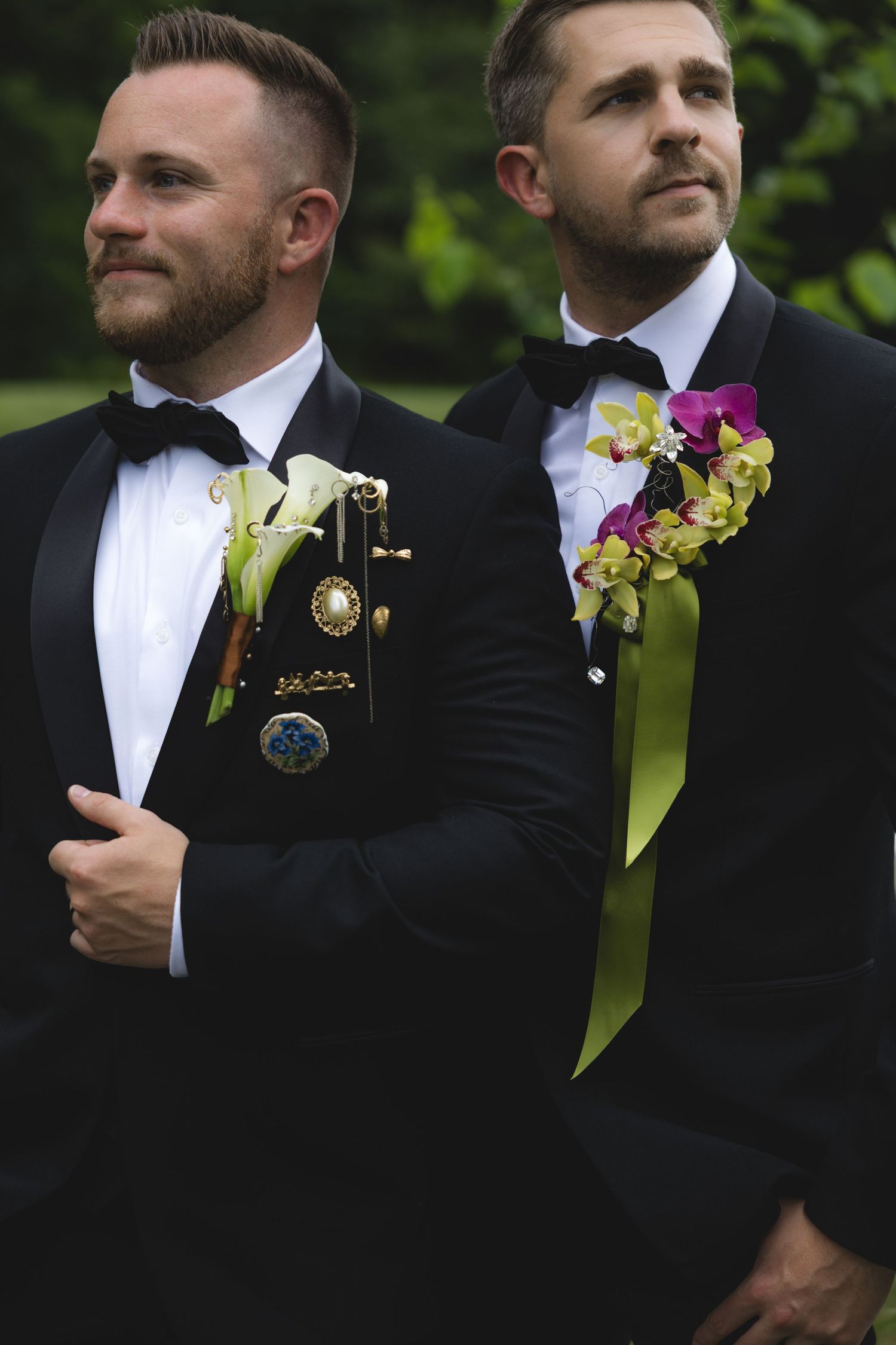 Two grooms in black tuxedos with floral boutonnieres pose outdoors on their wedding day.