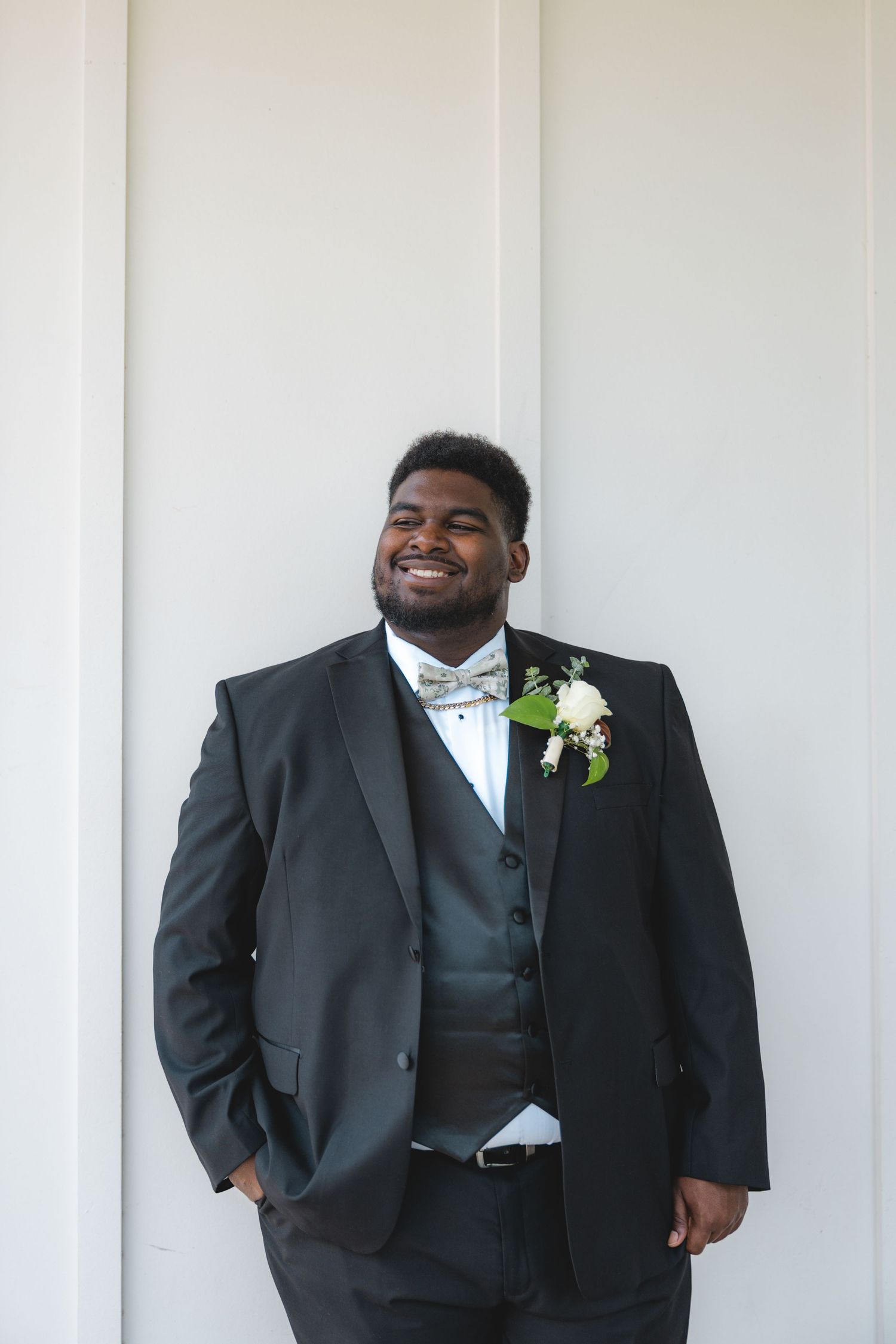 Handsome groom in black tuxedo with floral boutonniere smiles confidently against a white paneled wall.