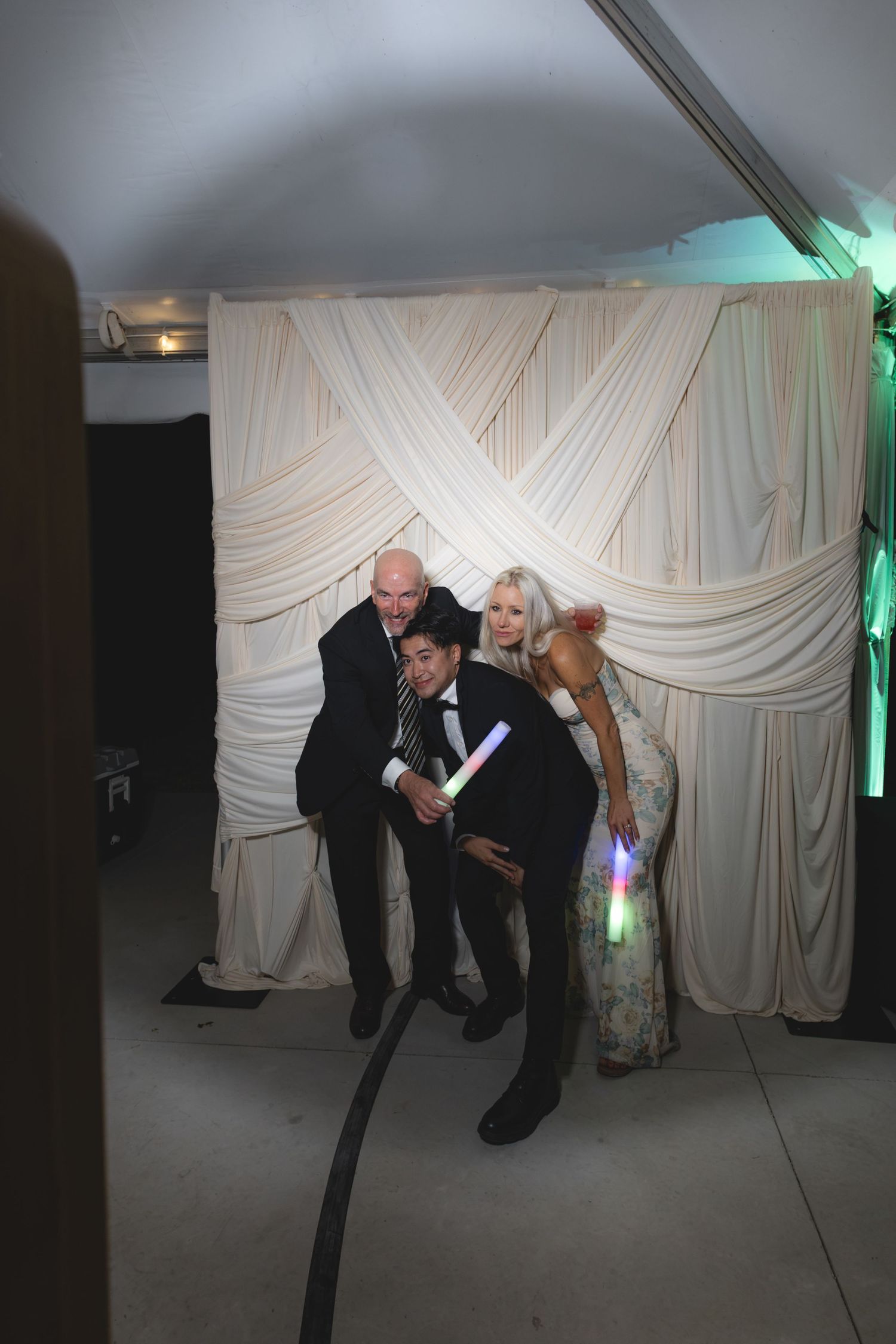 Three wedding guests posing playfully with glow sticks in front of draped backdrop.