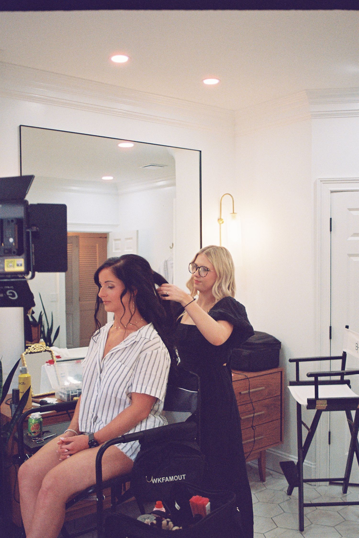 Blonde hairstylist styling brunette bride's hair in bright white salon before wedding day.