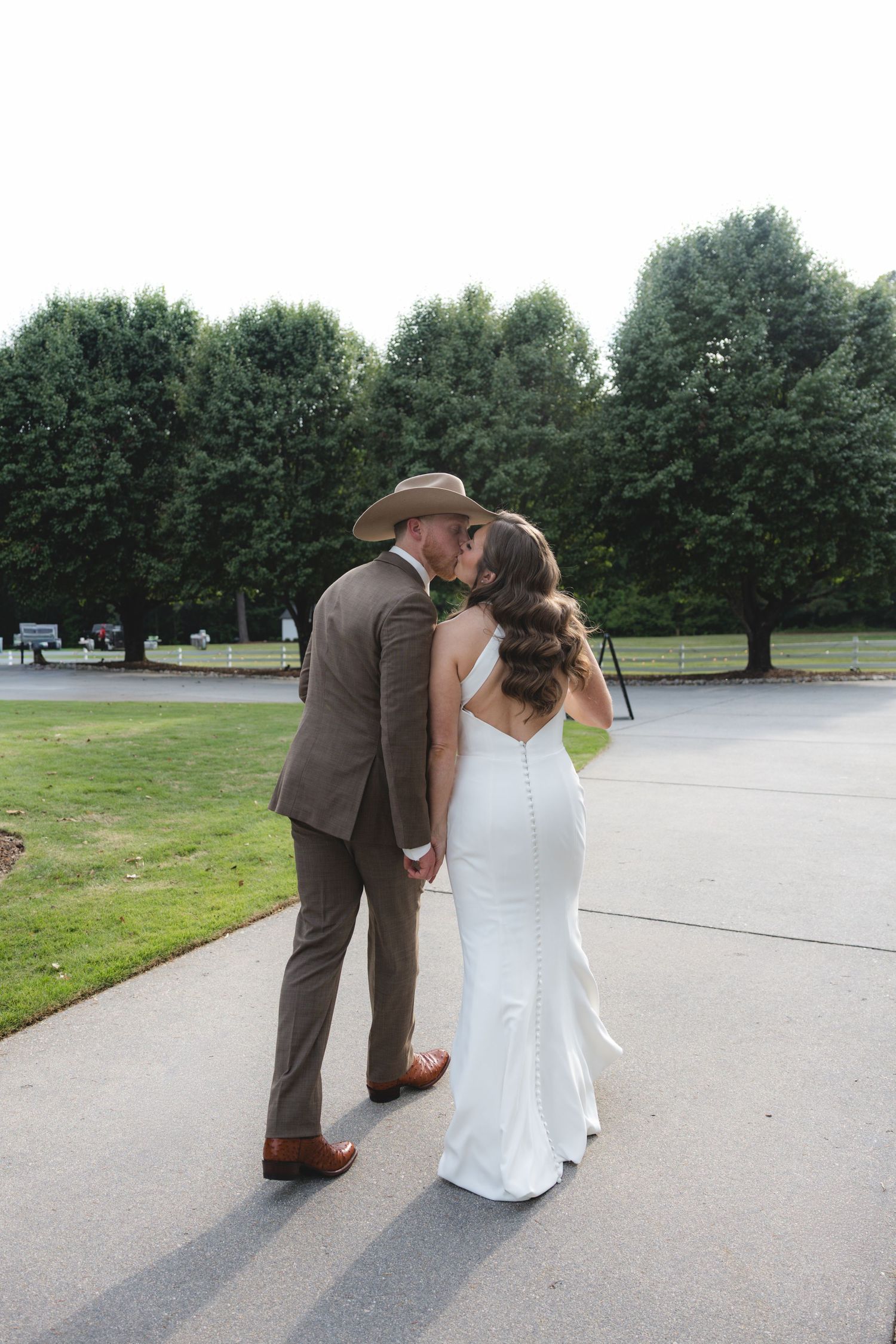 Bride in fitted white gown and groom in brown suit share romantic kiss outdoors.