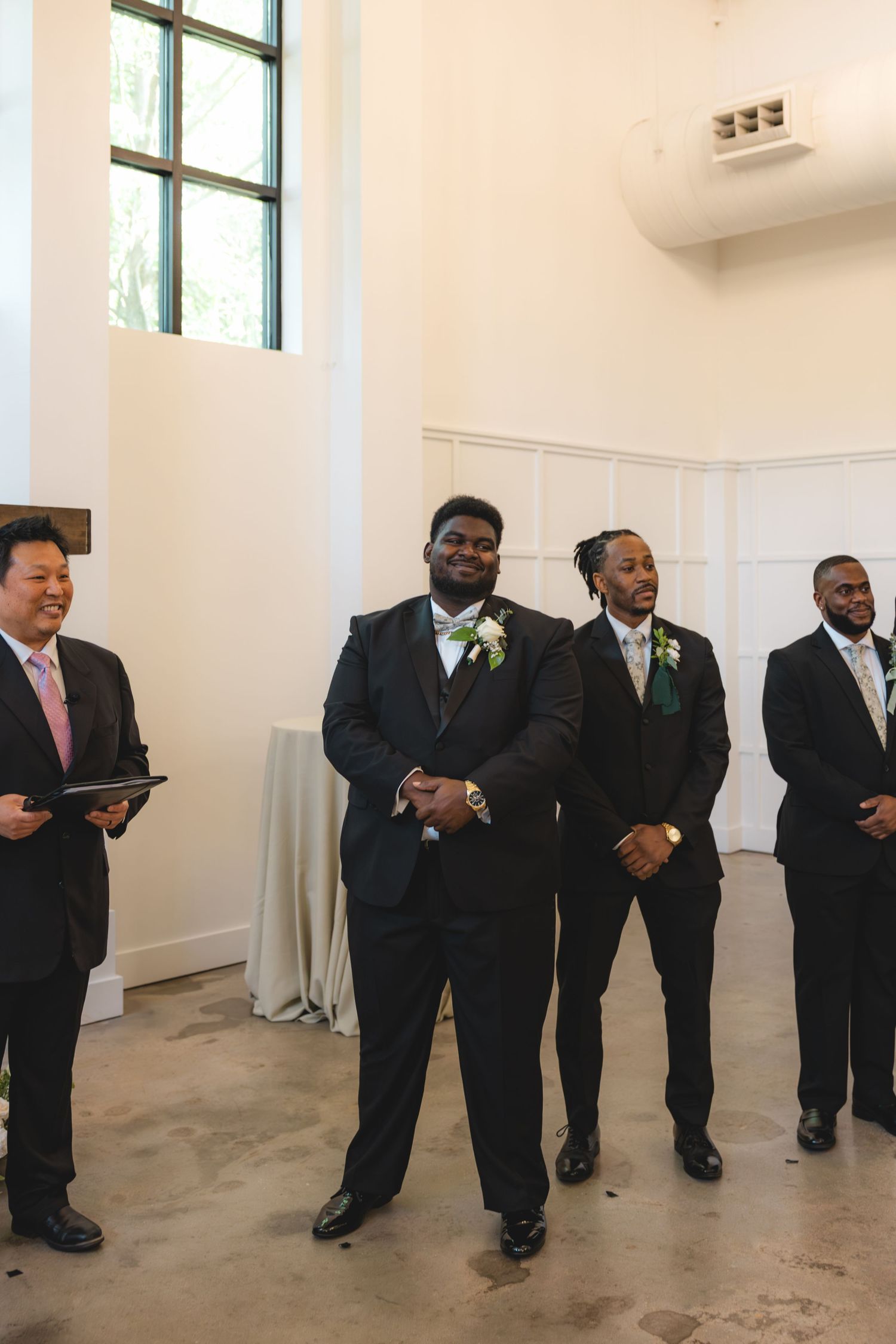 Groomsmen in black suits with boutonnieres stand at the wedding altar beside an officiant holding a book.