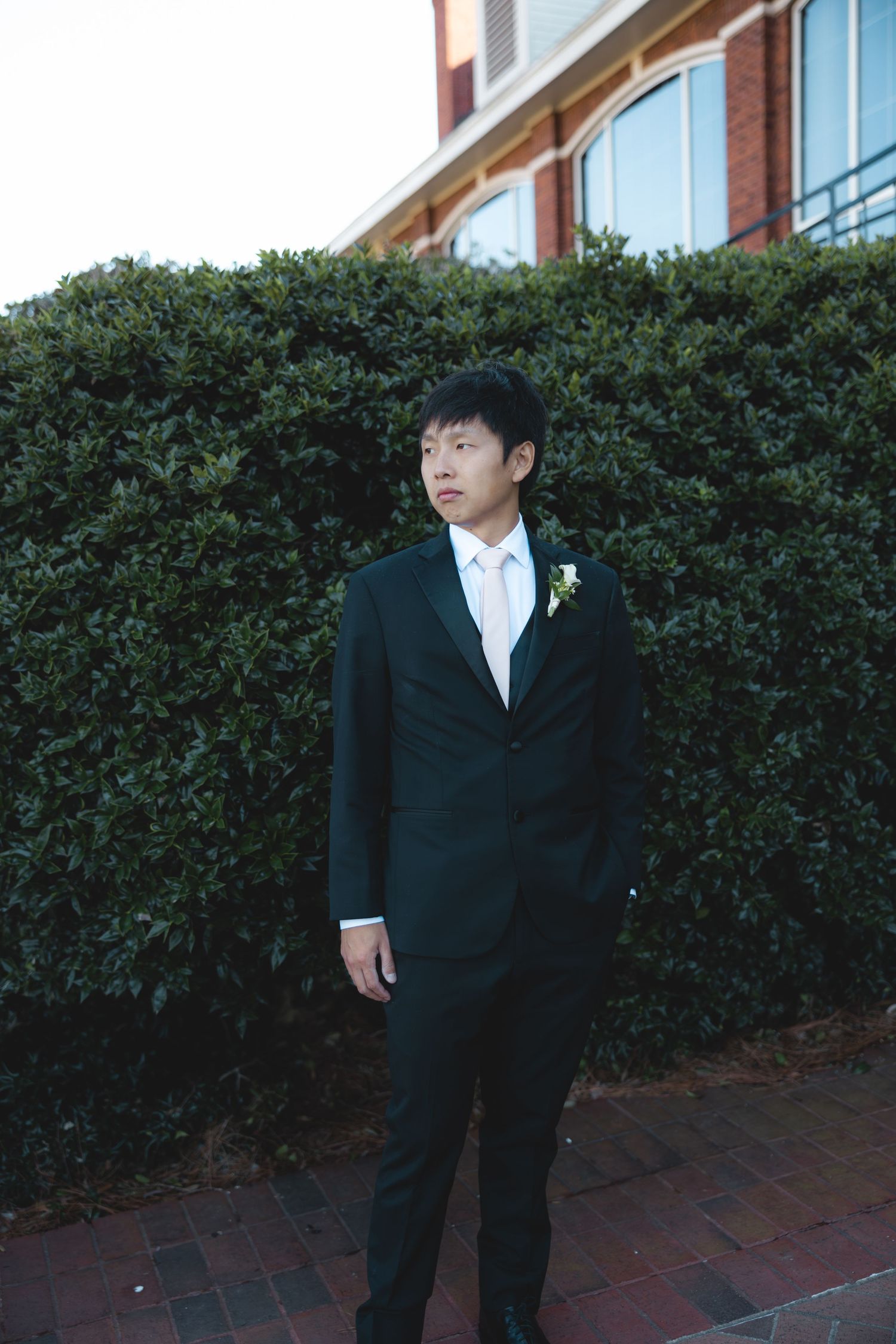 Handsome groom in black tuxedo with boutonniere standing beside lush green hedge.