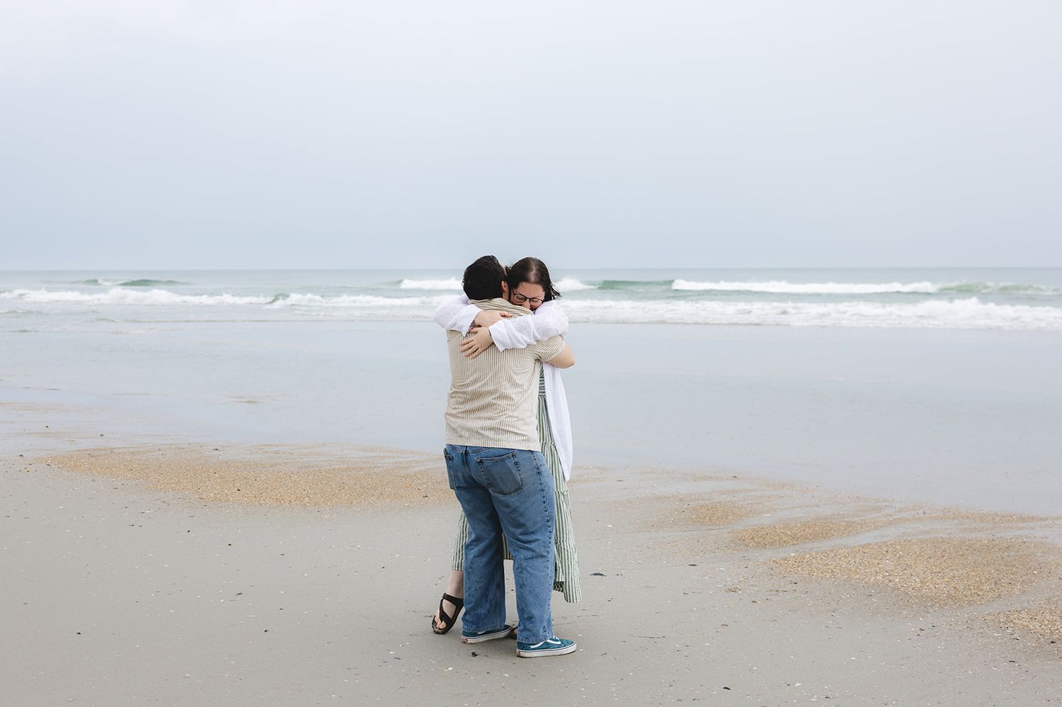 Two people embracing on a misty beach with ocean waves in the background on an overcast day.