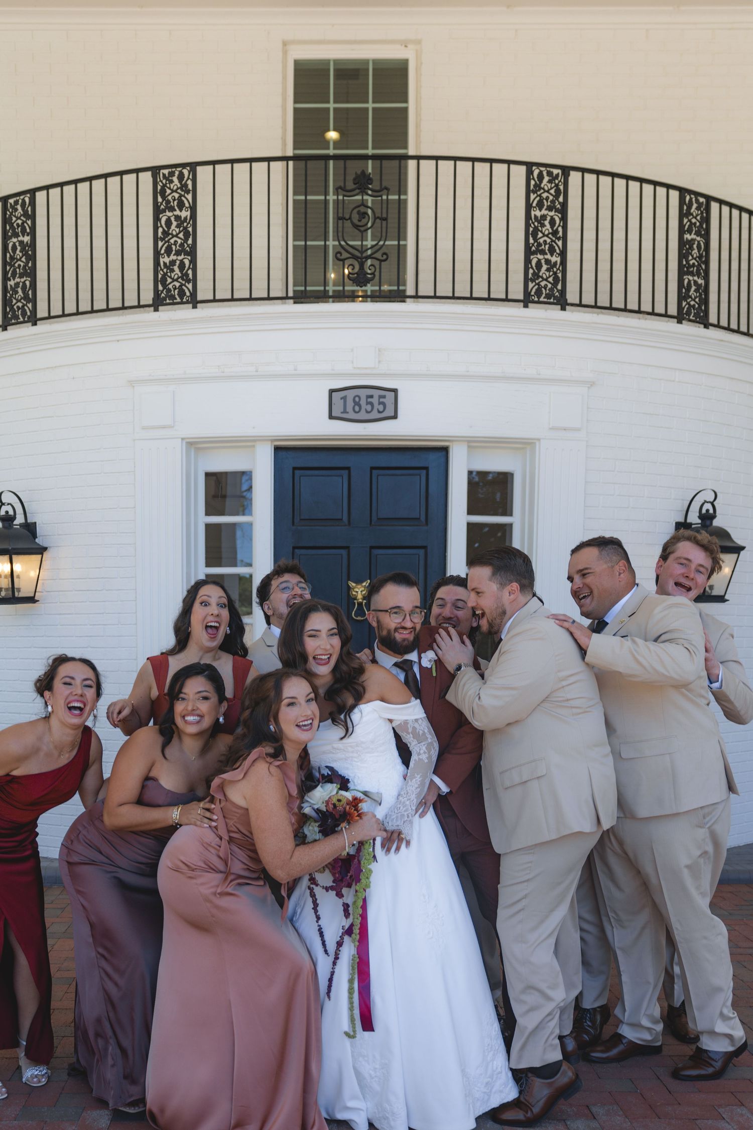 Wedding party celebrating outside a white building with balcony, bride in white gown surrounded by bridesmaids and groomsmen.