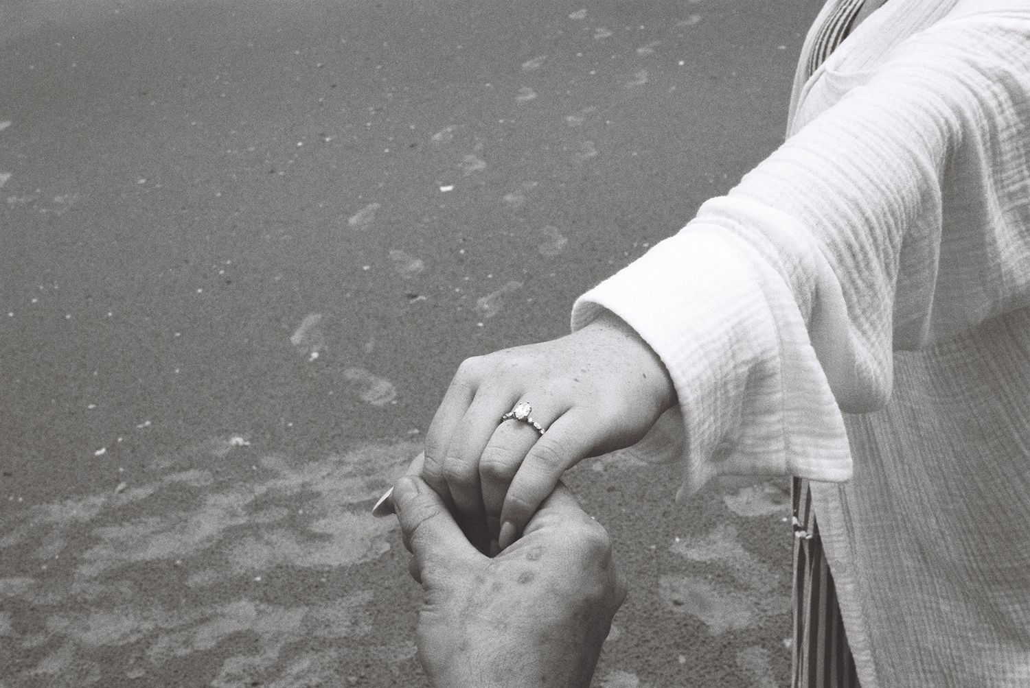 Black and white photo of two hands holding, one wearing a diamond ring, near a sandy beach.