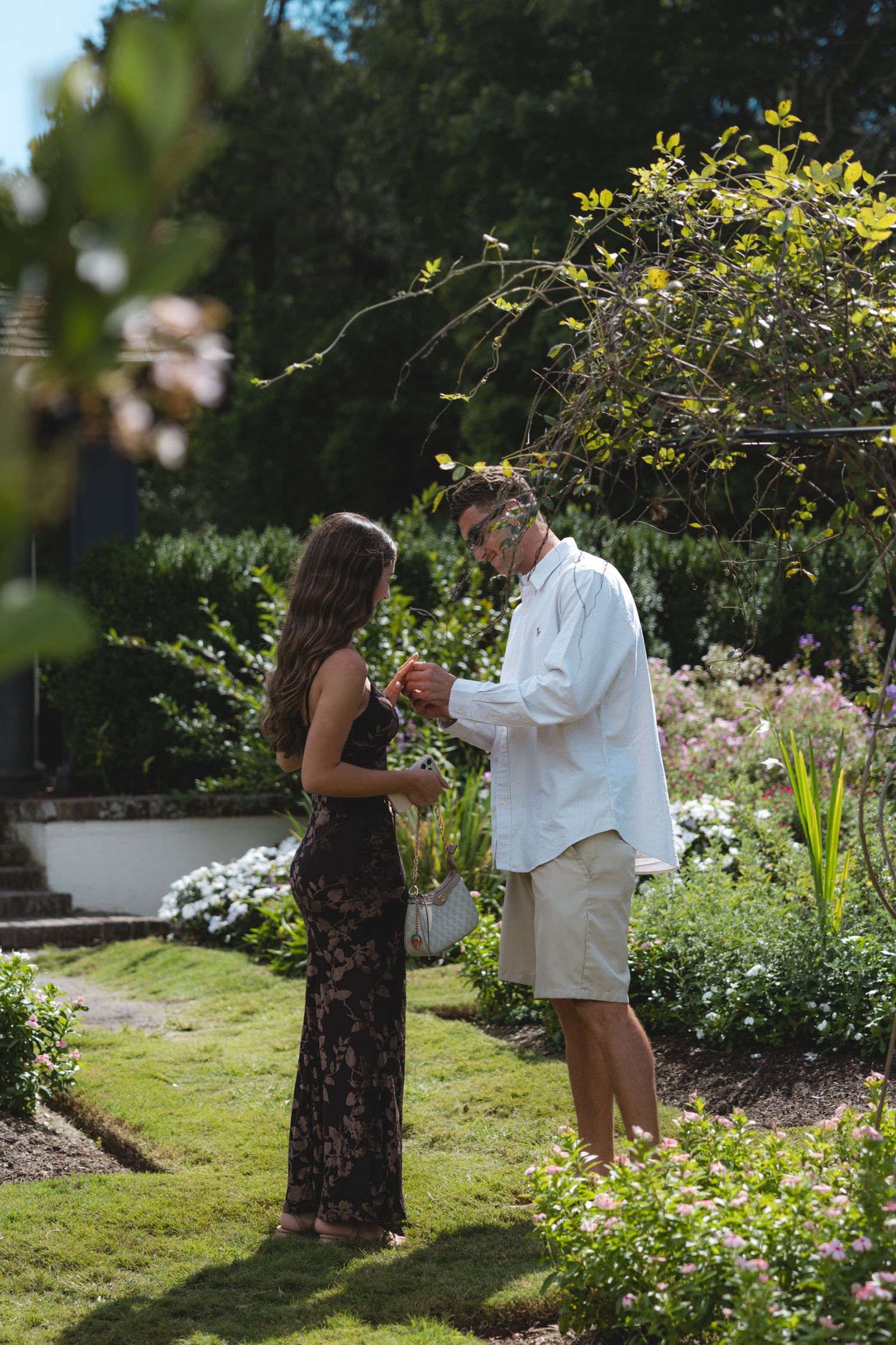 Couple standing in a lush garden on a sunny day, woman in floral dress and man in white shirt exchanging a flower.