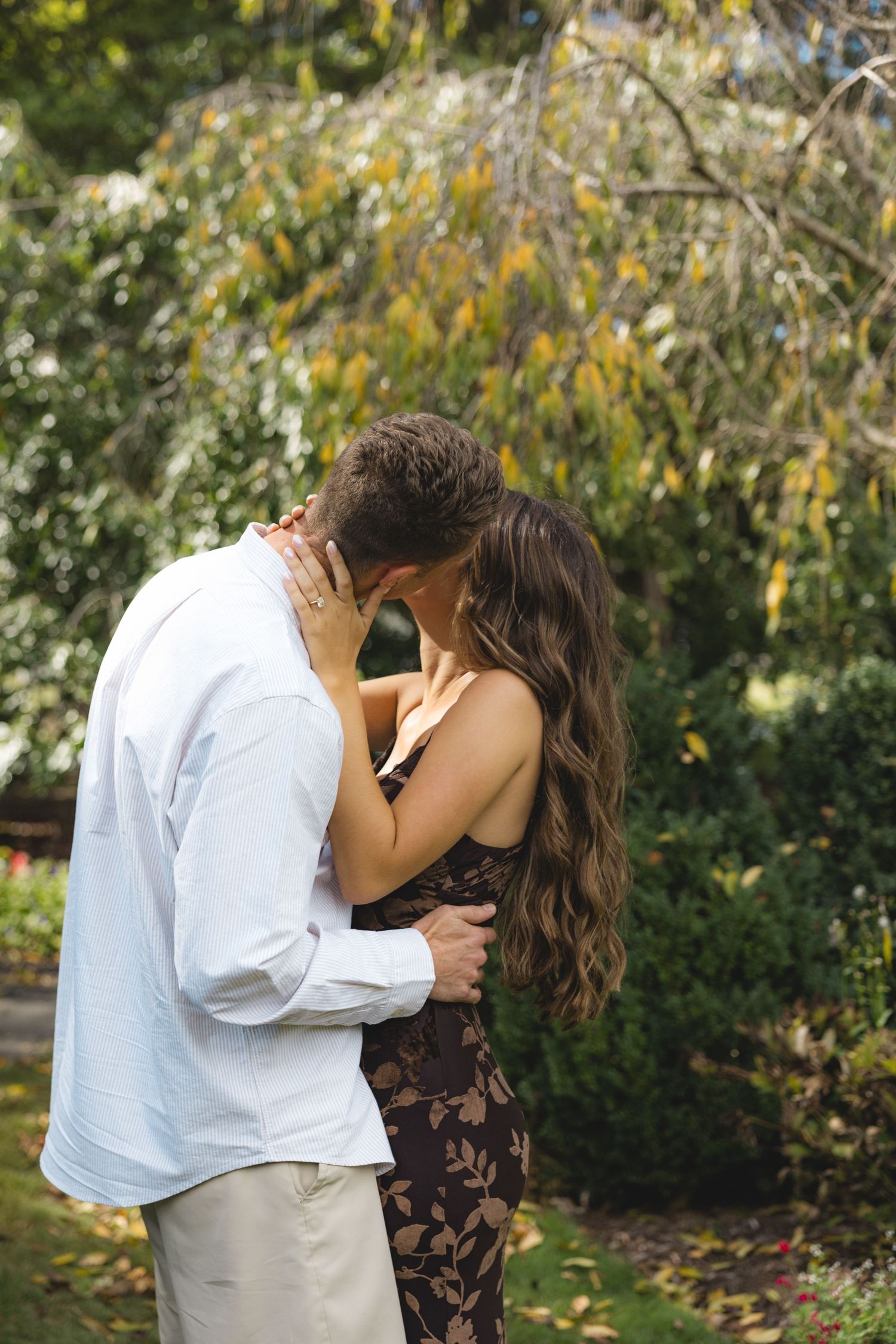 Couple sharing a romantic kiss in a lush garden, woman in floral dress with long wavy hair, man in white shirt.