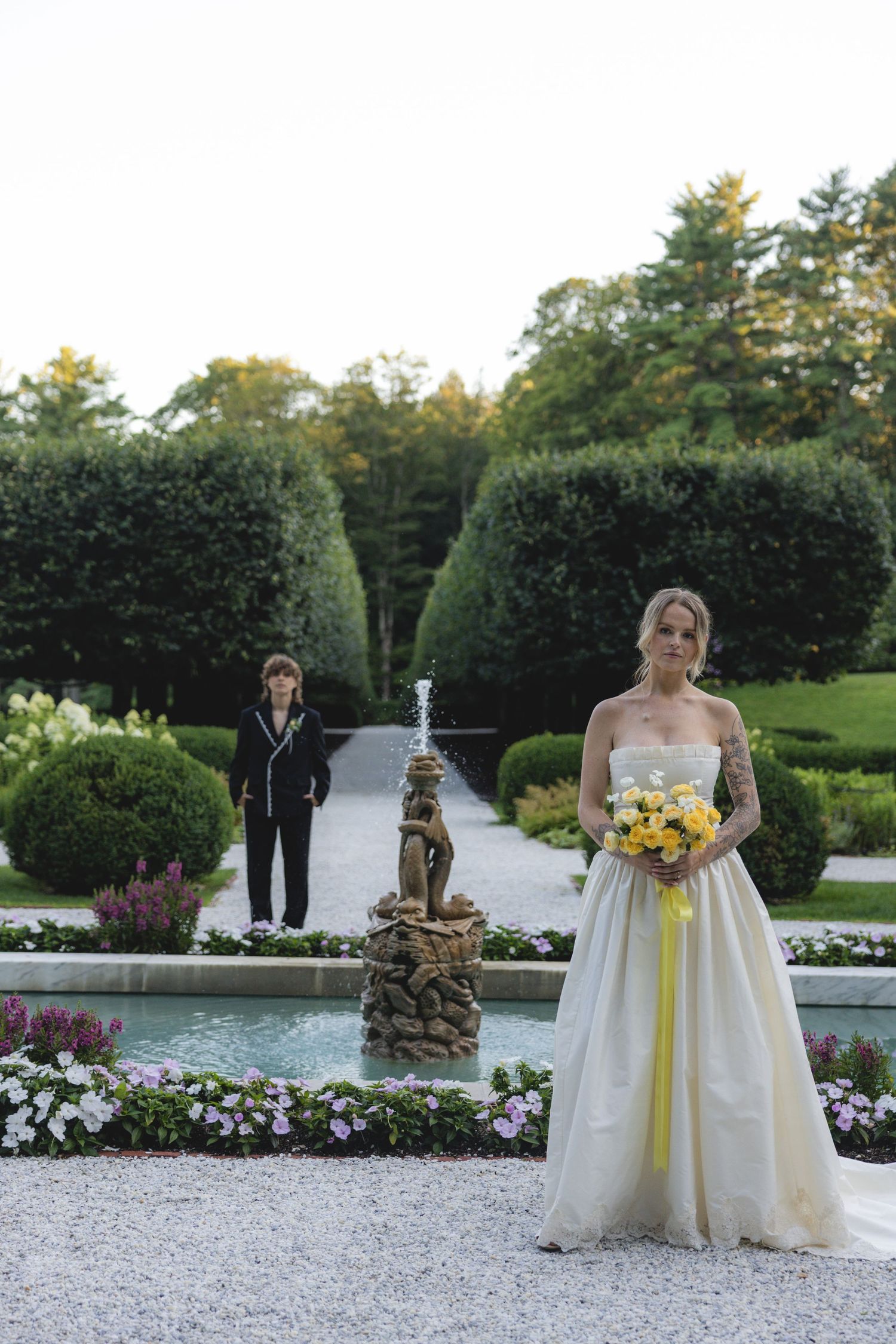 Bride in strapless gown holding yellow bouquet stands before garden fountain with wedding guest in background.