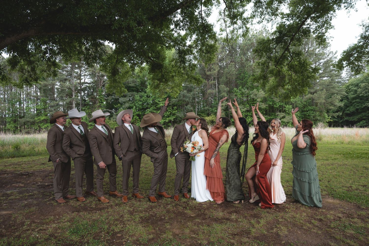 Wedding party in rustic outdoor setting, groomsmen in brown suits and bridesmaids in earthy tones celebrating under large tree.