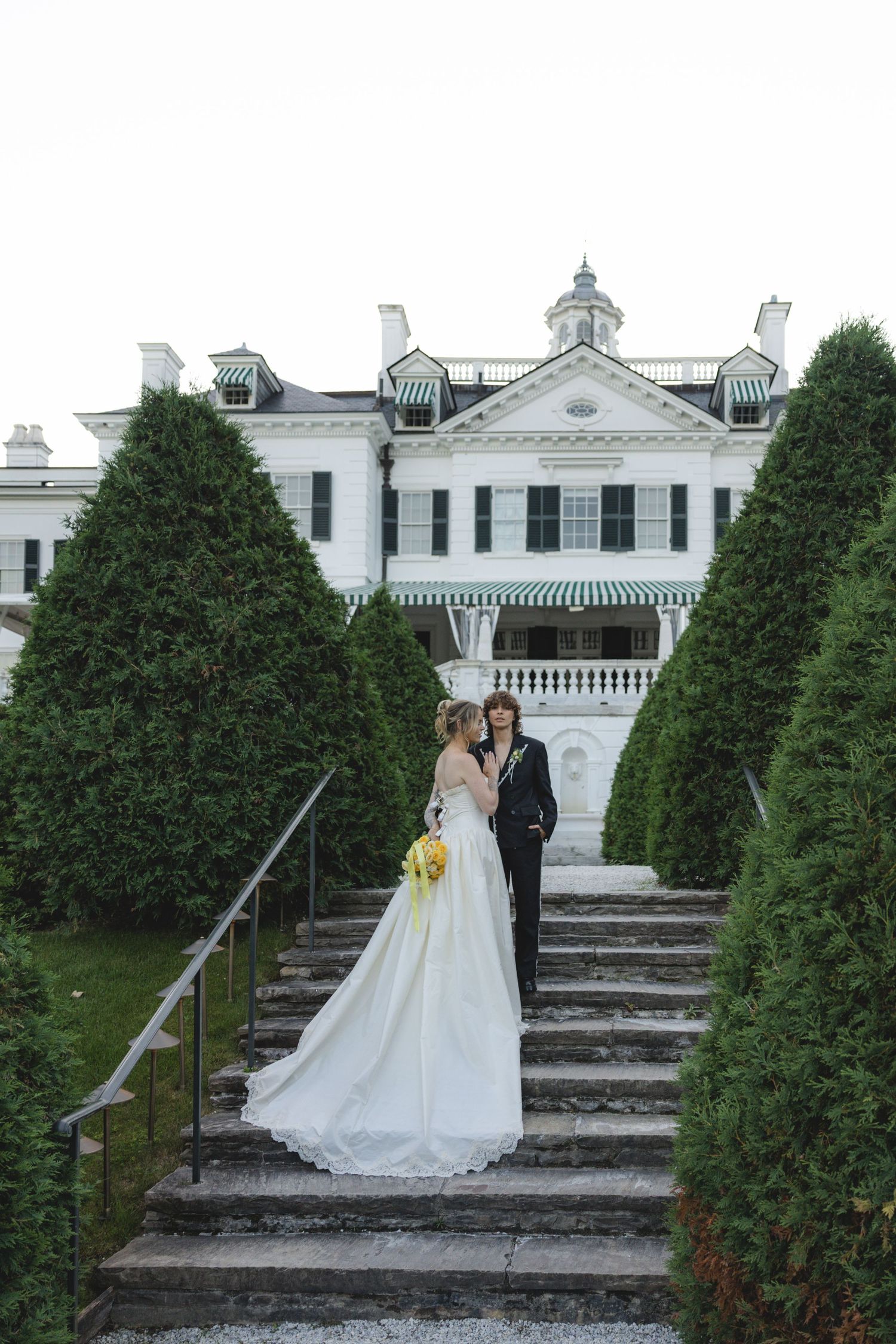 Bride and groom pose on garden steps before a grand white colonial mansion surrounded by manicured evergreen trees.