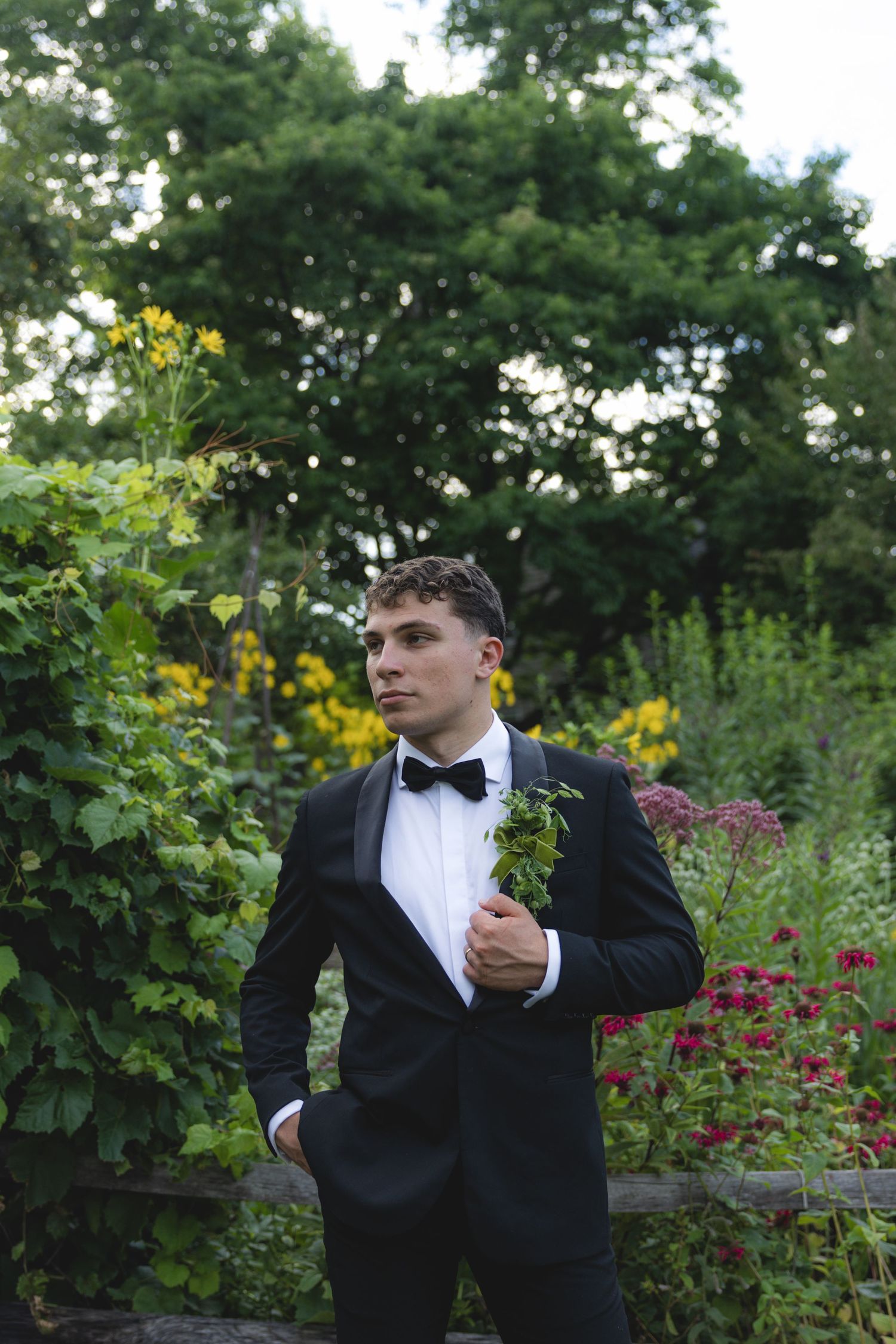 Young man in black tuxedo with bow tie and boutonniere poses in lush garden with colorful flowers.