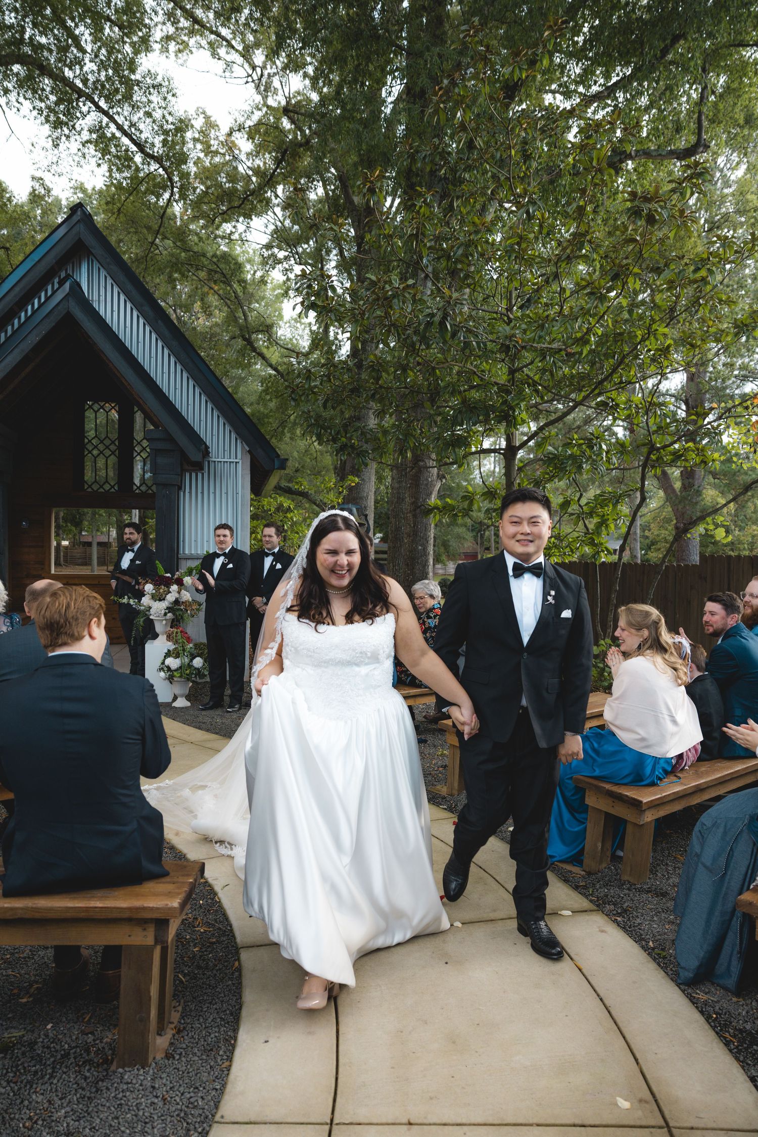 Smiling bride in a white gown and groom in a black suit hold hands walking down the aisle past seated guests at an outdoor wedding.