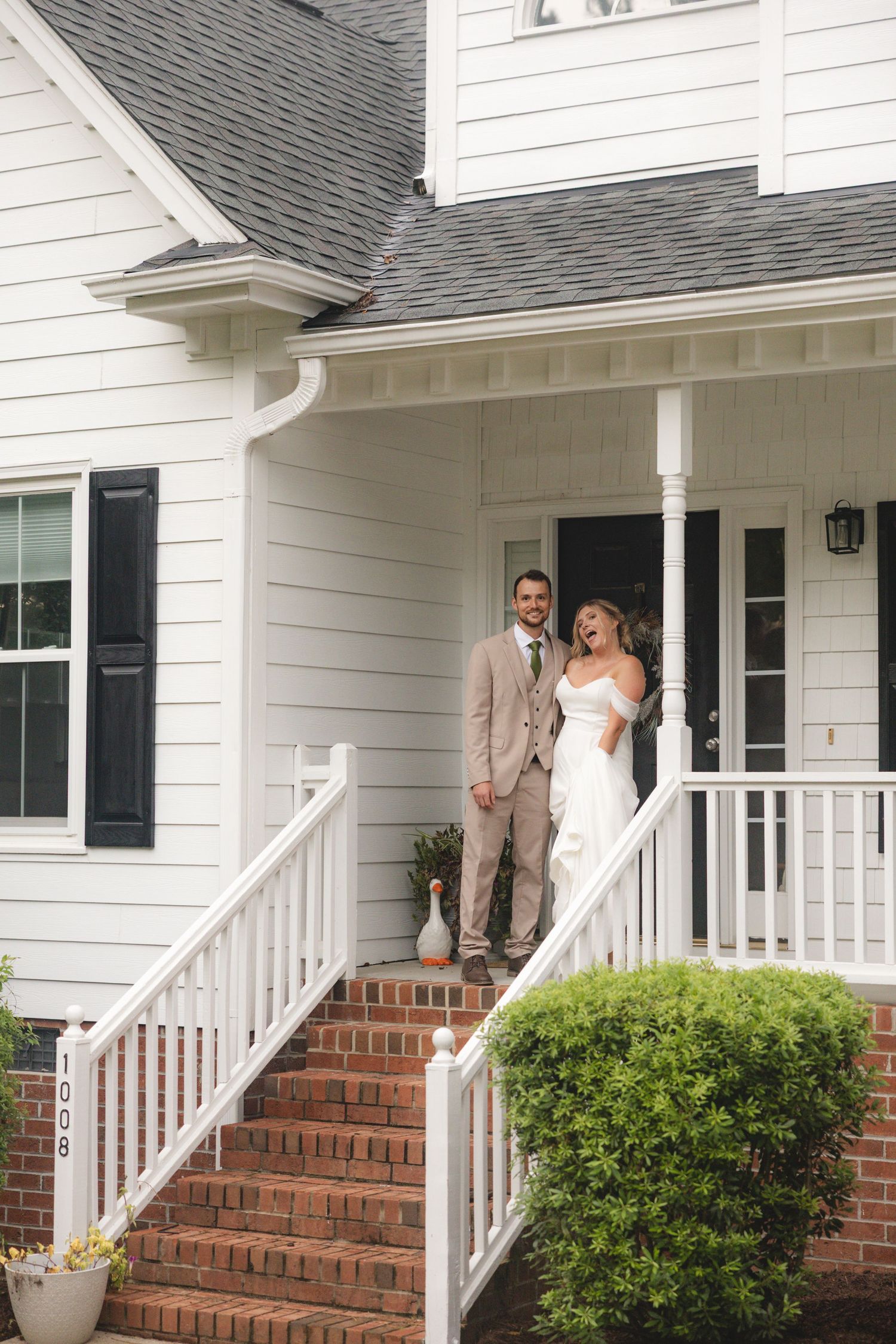 Bride and groom smiling together on brick porch steps of white farmhouse home on their wedding day.
