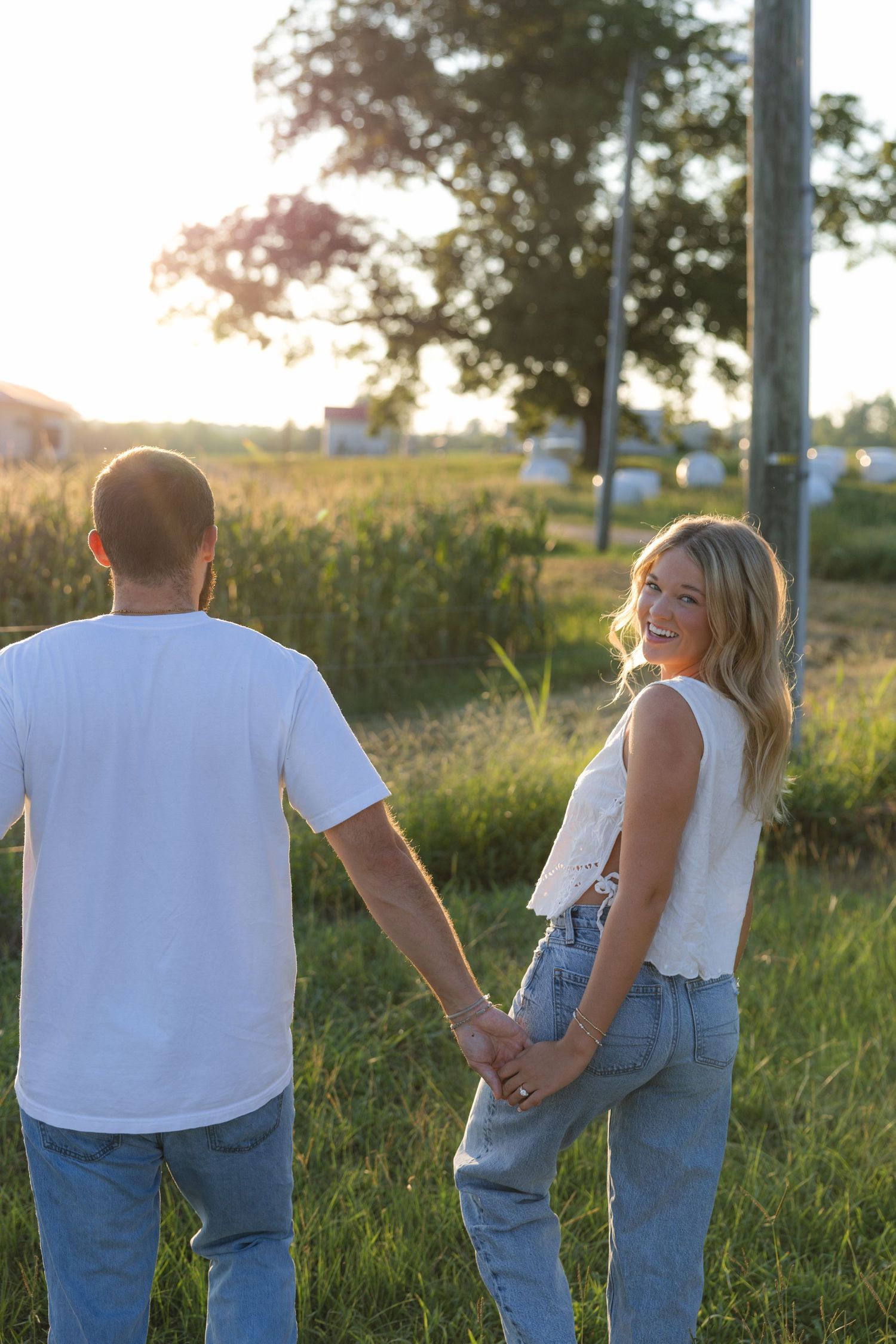 A smiling blonde woman holds hands with a man in a white tee, walking through a sunny green field at golden hour.
