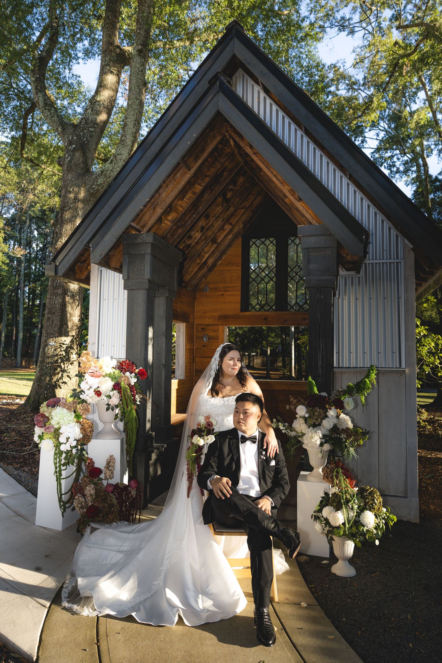 Bride in a white gown and groom in a black tuxedo pose on a bench outside a rustic wooden chapel with elegant floral arrangements.