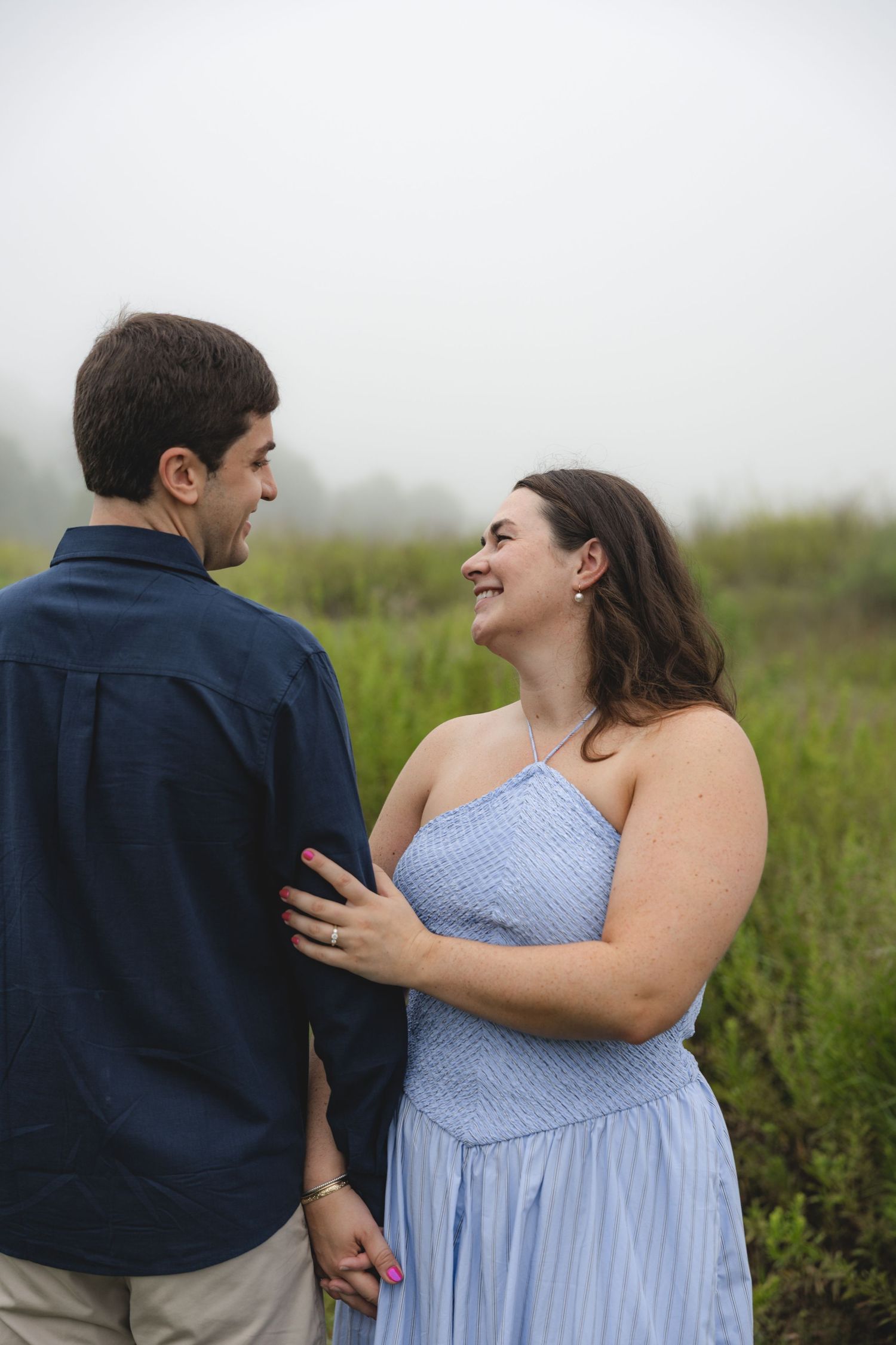 Couple smiling at each other in a misty field, woman in blue dress holding man in navy shirt.
