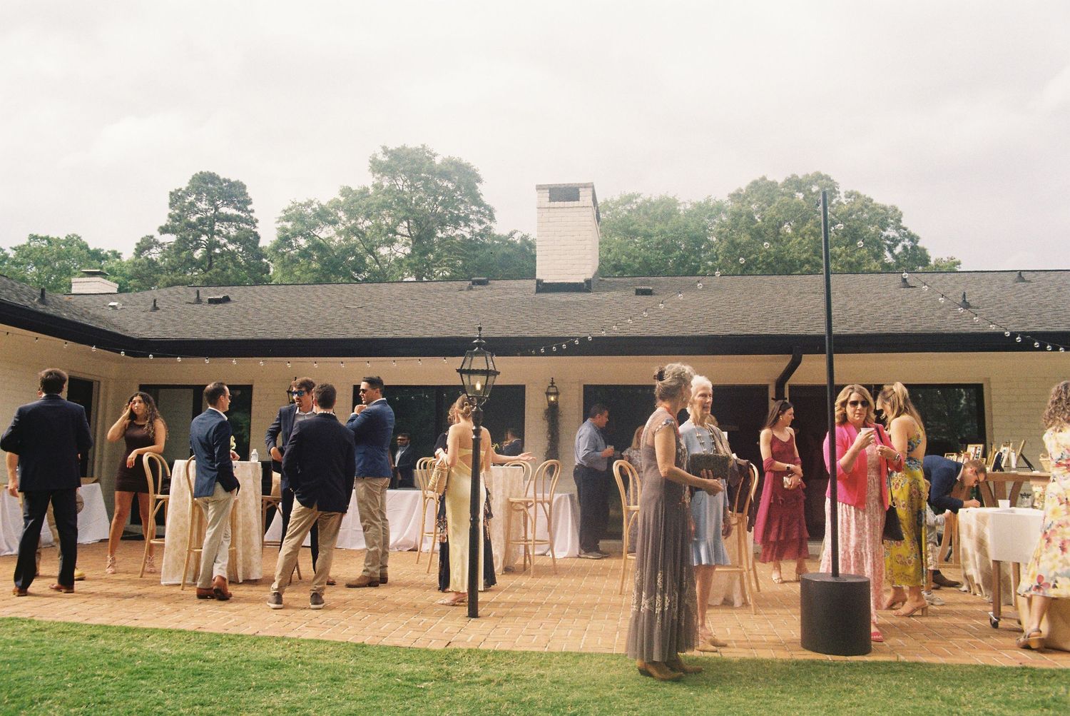 Guests mingle outdoors at an elegant garden wedding reception beside a white single-story venue surrounded by lush trees.