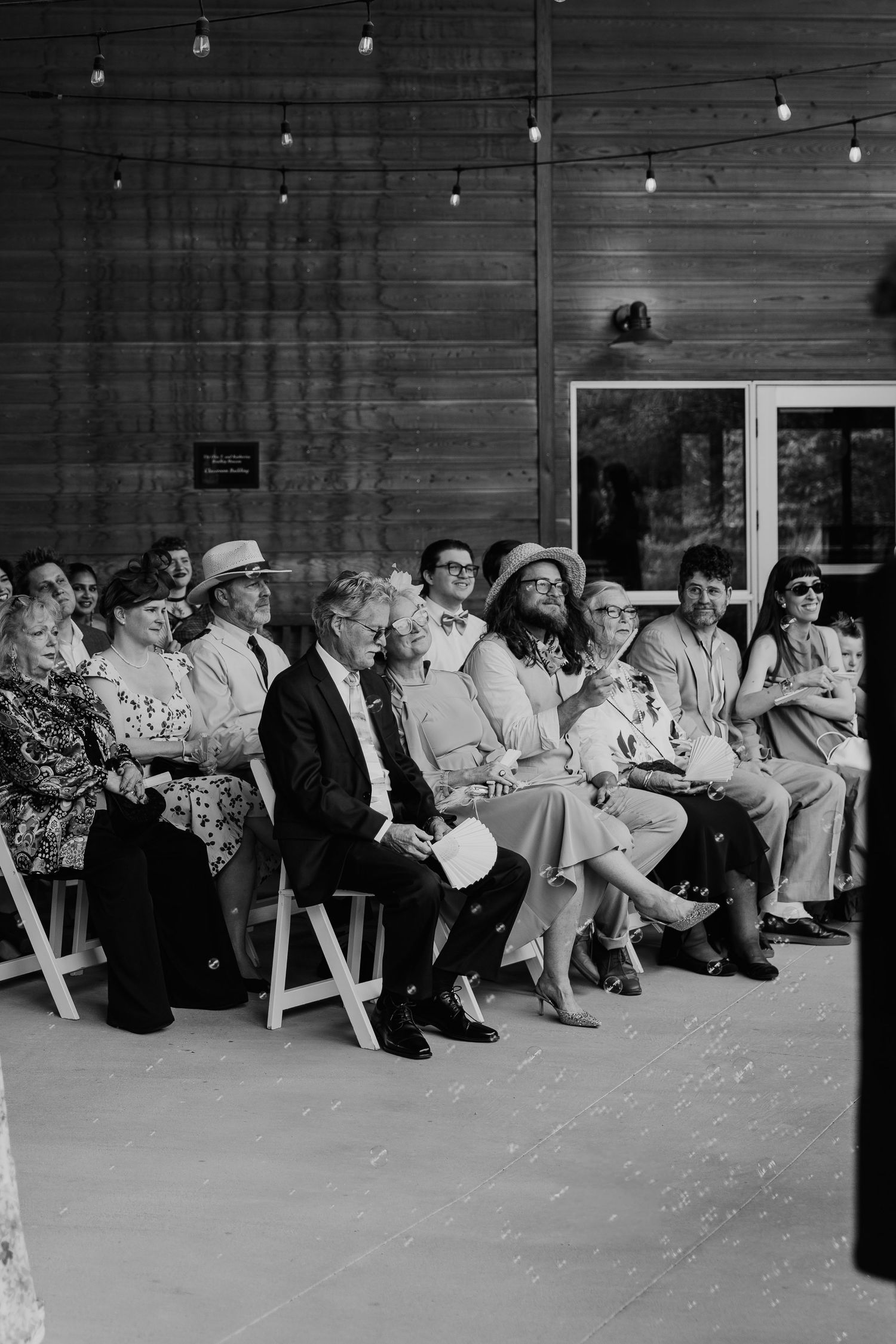 Black and white photo of wedding guests seated in chairs, smiling and watching a ceremony at a rustic wood-paneled venue.