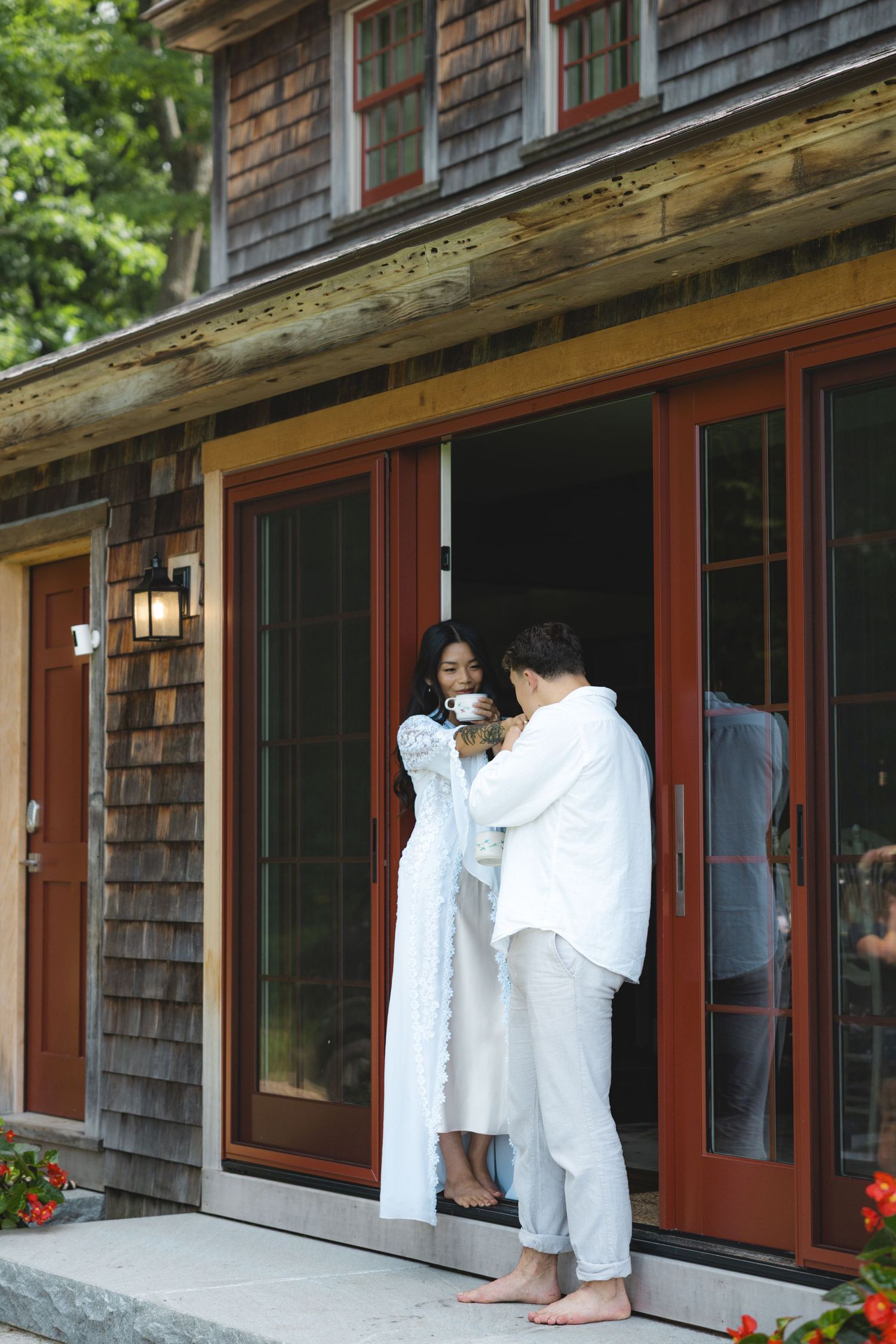 Couple in white robes sharing a morning coffee outside a rustic cedar shingle home with red French doors.