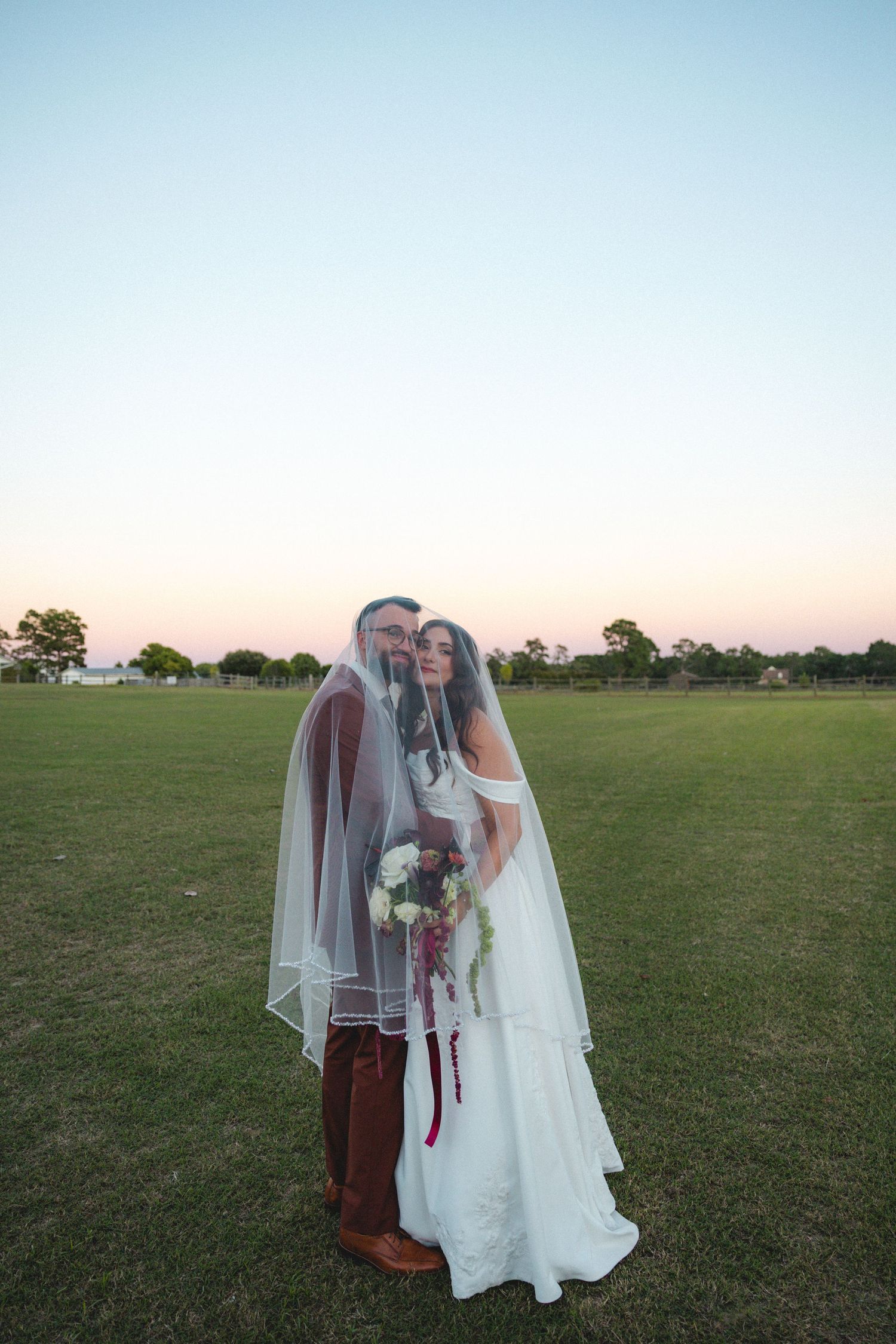 Bride and groom share a romantic moment under a veil at sunset on a green field, bride holding a bouquet.