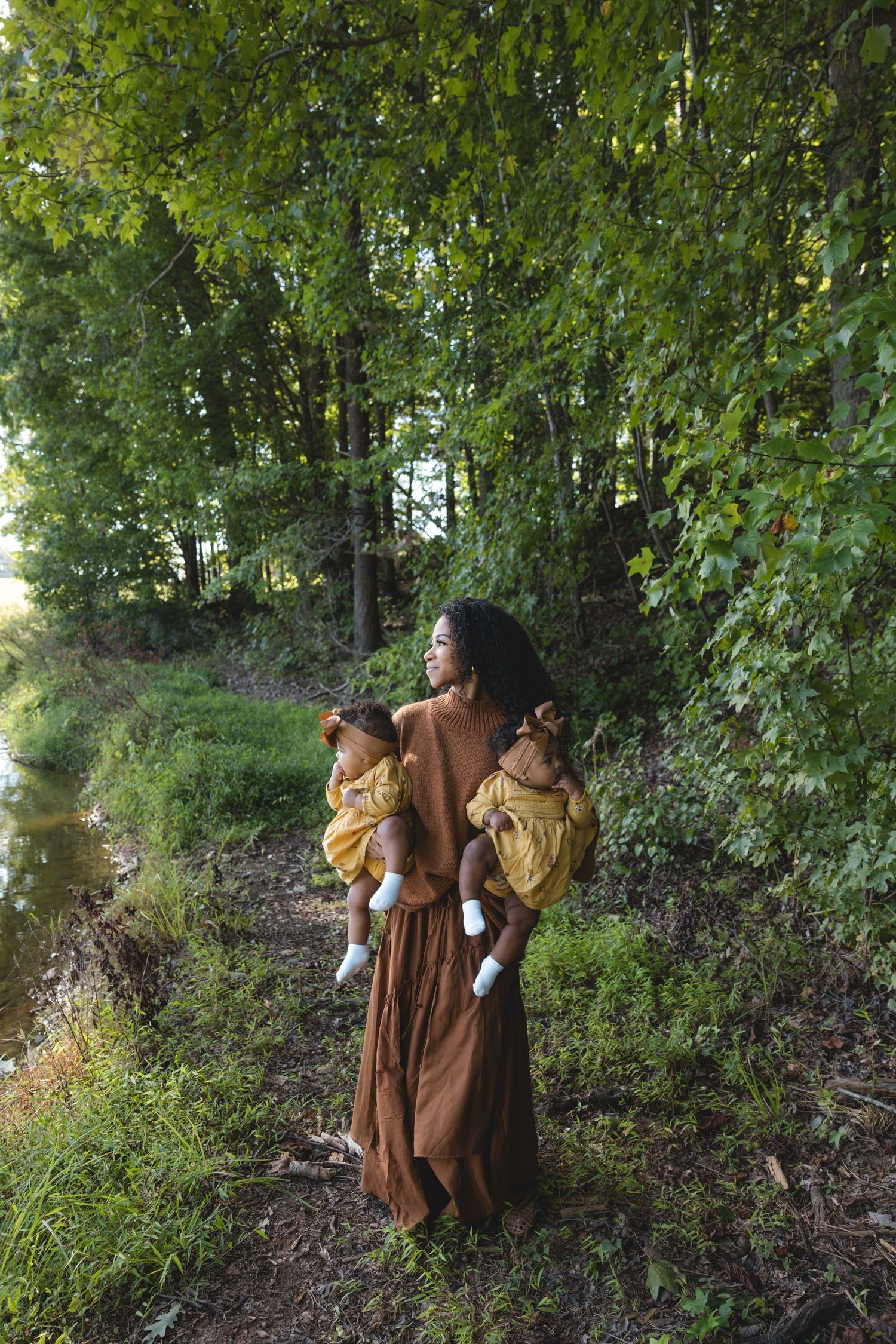 A woman in a brown dress holds twin babies in yellow outfits while standing on a forest path near a stream.