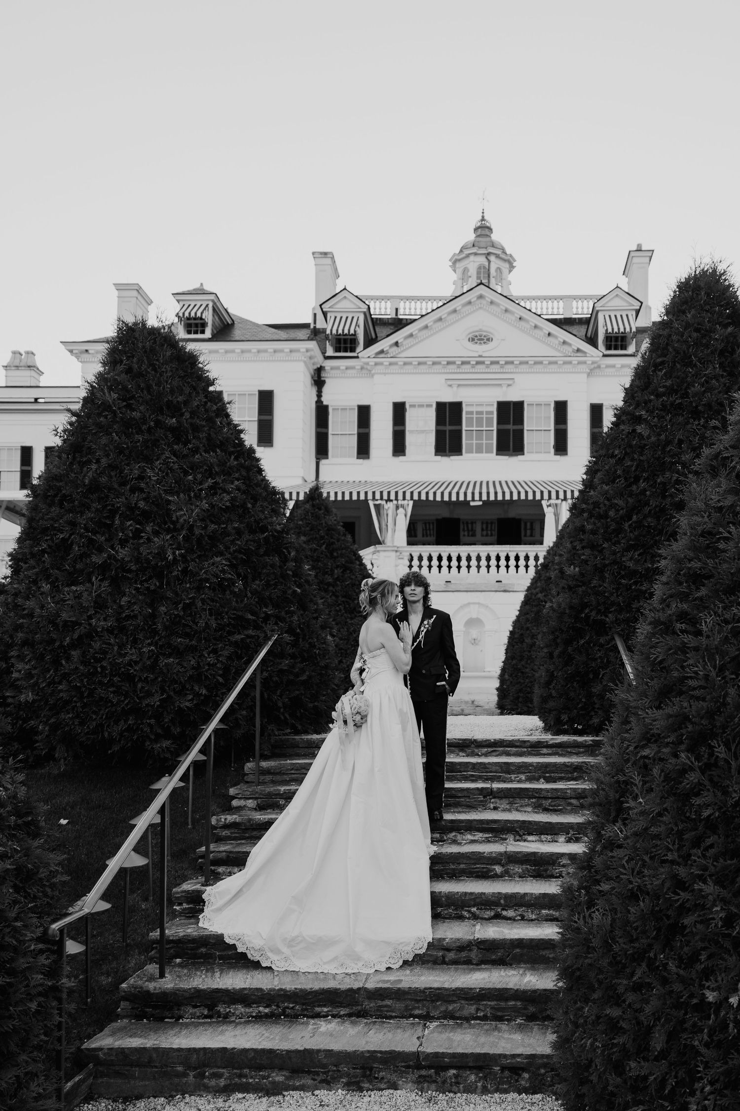 Black and white photo of a bride and groom posing on garden steps before a grand estate mansion.