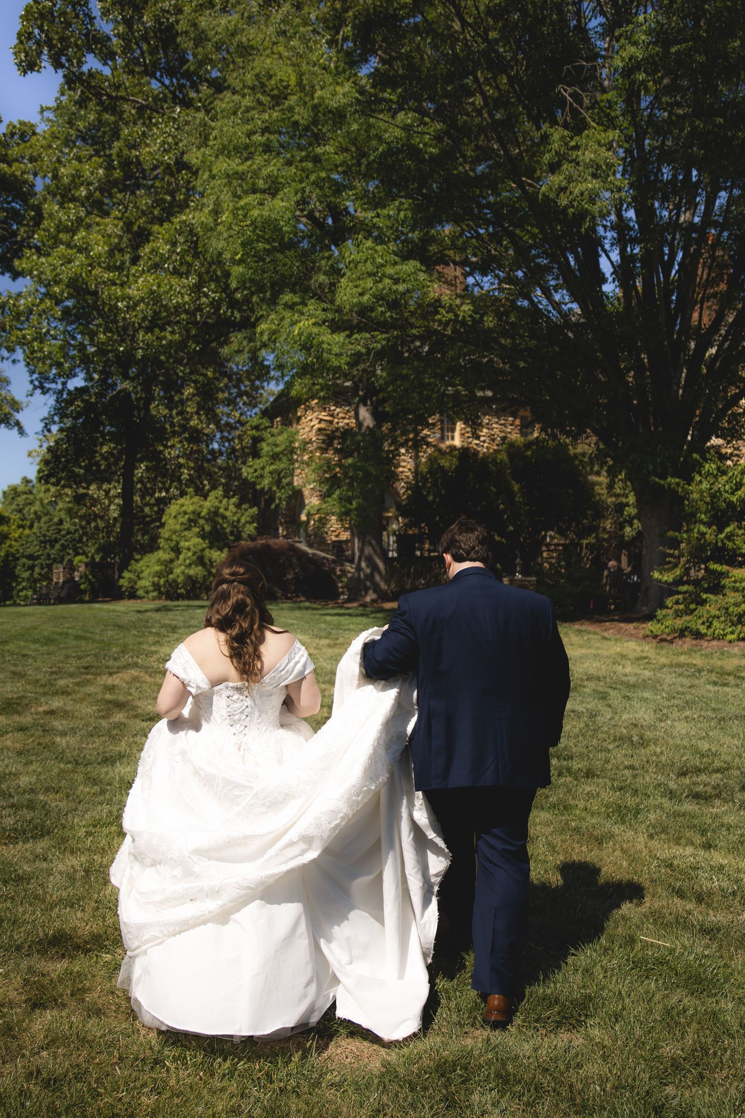 Bride and groom walk hand in hand across a green lawn surrounded by tall trees on their wedding day.