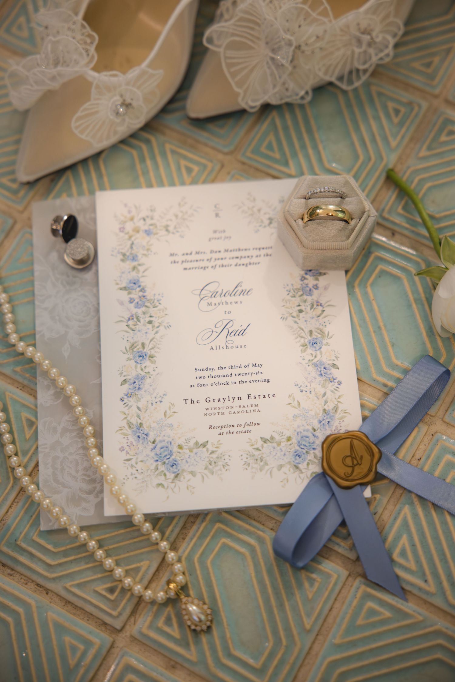 Elegant wedding invitation with blue floral border, pearl necklace, gold wax seal, rings, and bridal heels on tiled surface.