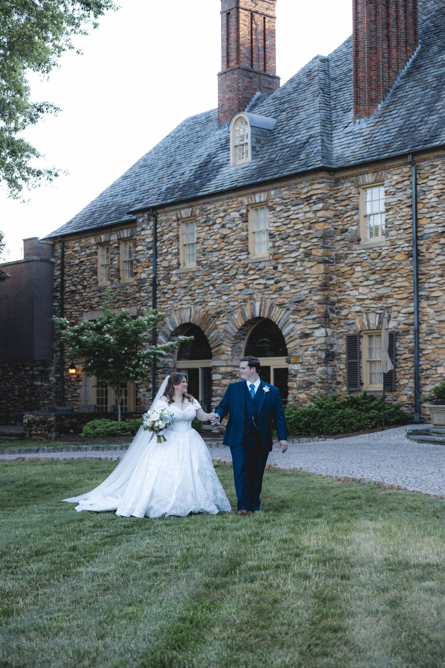 Bride and groom walk hand-in-hand on green lawn before a stone mansion at dusk wedding.
