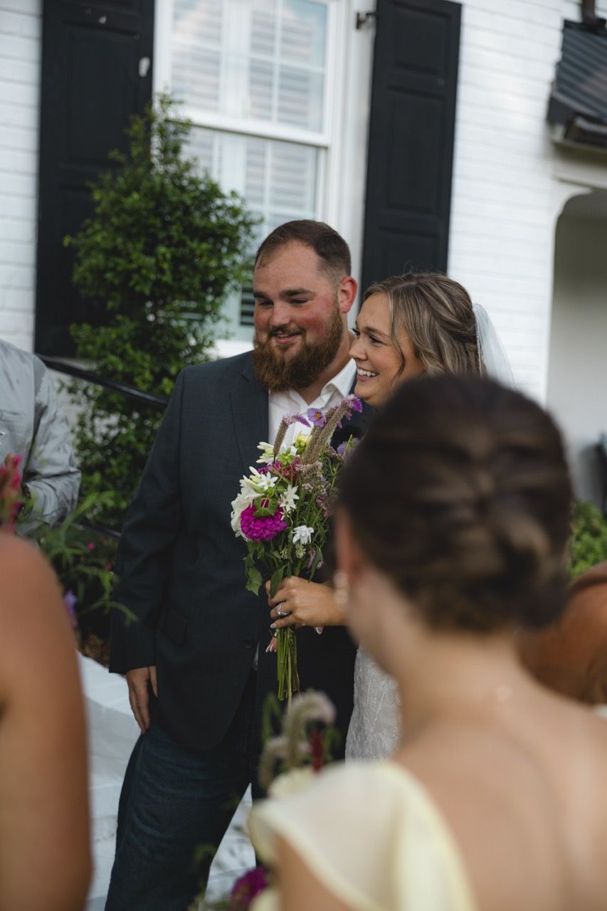 Smiling bride holding colorful bouquet walks with bearded groom in dark suit outside white house at outdoor wedding.