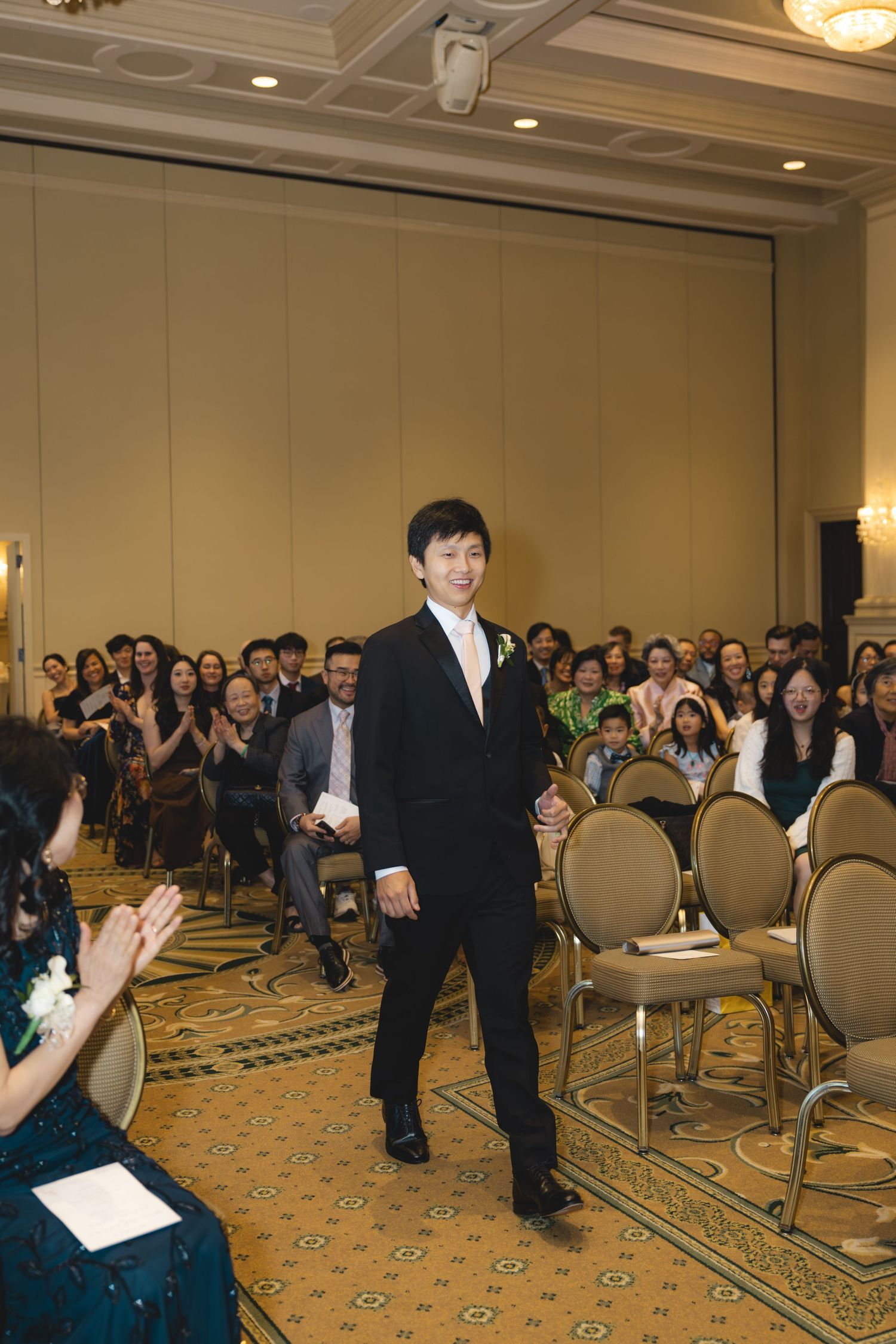 Groom in black tuxedo walks down aisle as wedding guests applaud in elegant ballroom.
