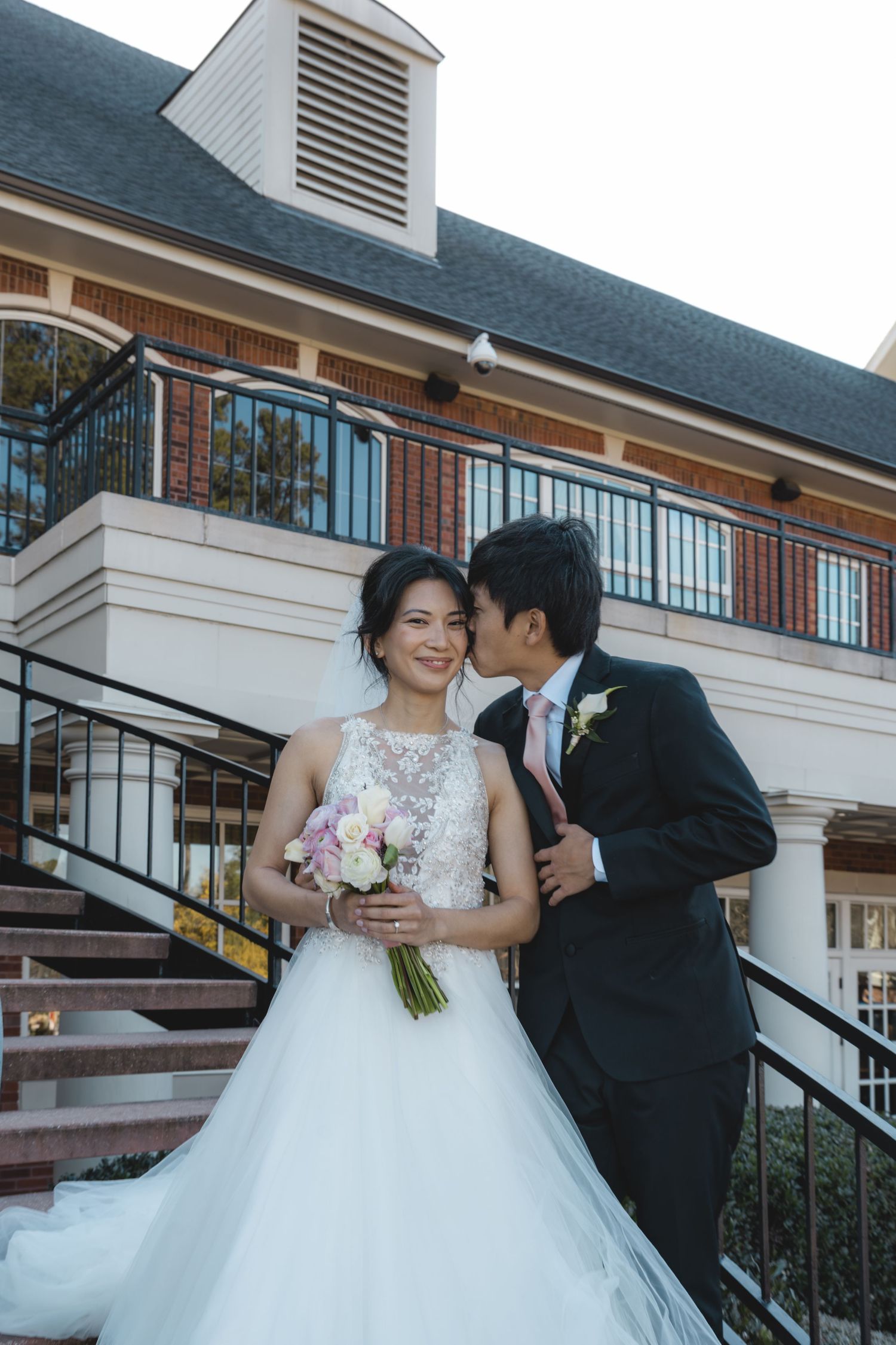 Groom kissing bride on cheek while she smiles holding bouquet on venue staircase.
