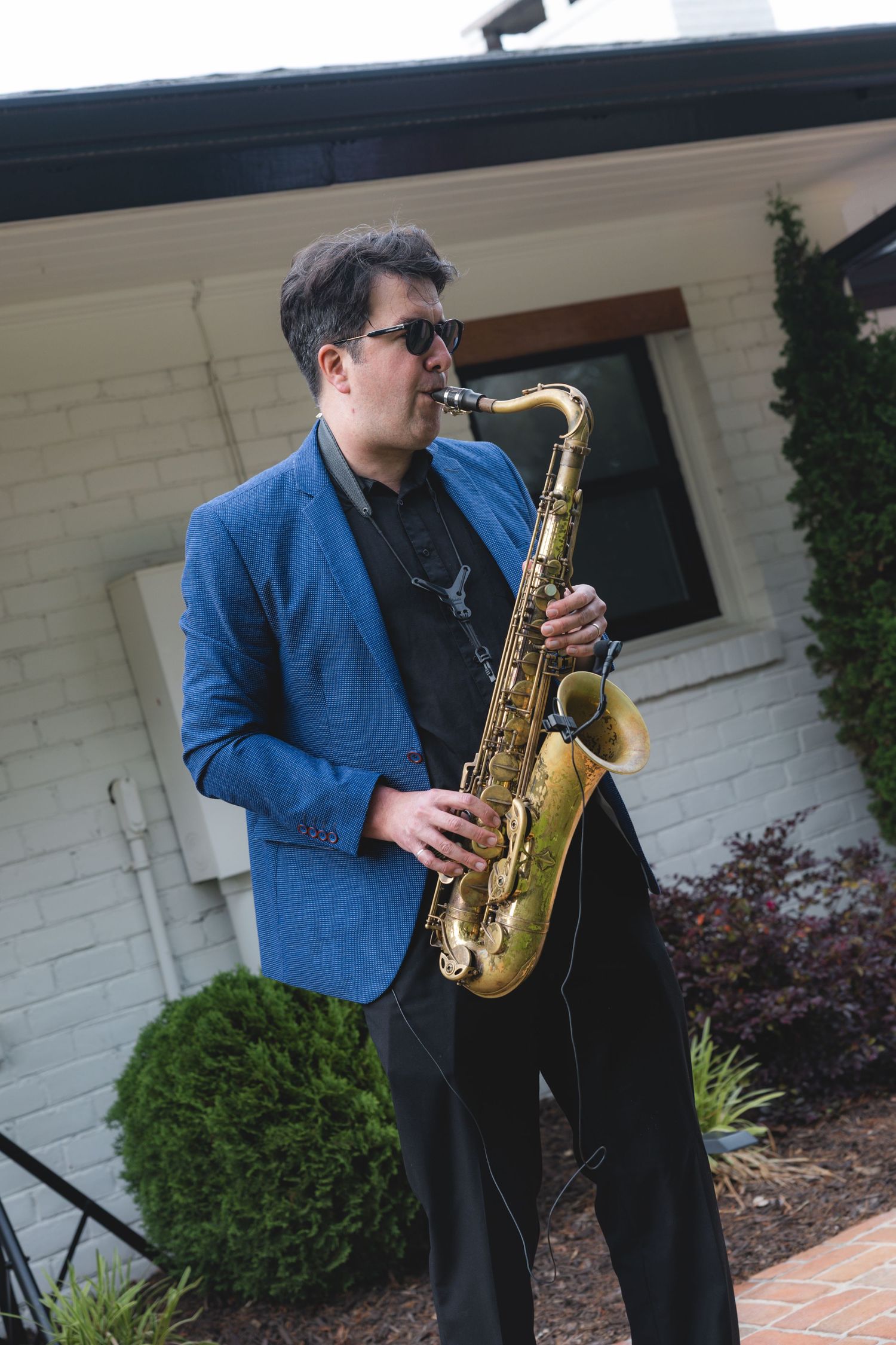 Musician in blue blazer plays saxophone outside white brick building at wedding reception.