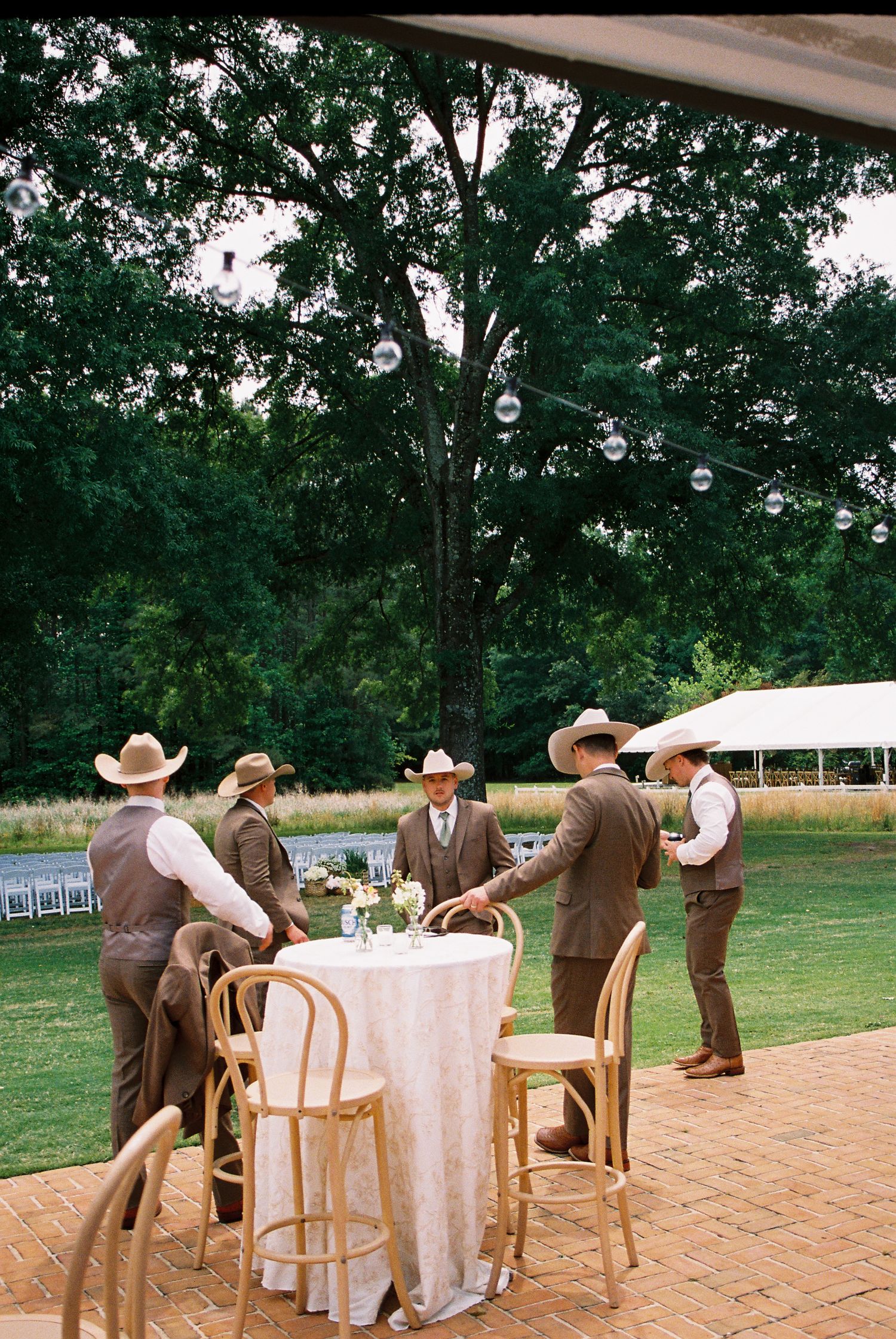 Groomsmen in brown suits and cowboy hats gathered around cocktail table at outdoor wedding reception.