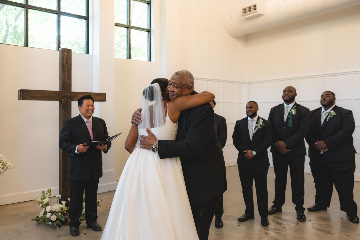 Bride and older man share warm hug at altar while groomsmen and officiant look on during ceremony.