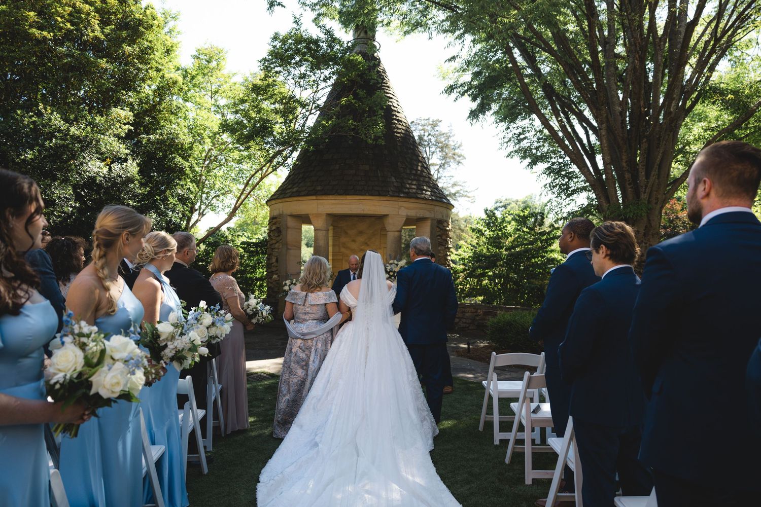 Bride walking down outdoor aisle in white gown with veil, flanked by bridesmaids in blue dresses at garden wedding ceremony.