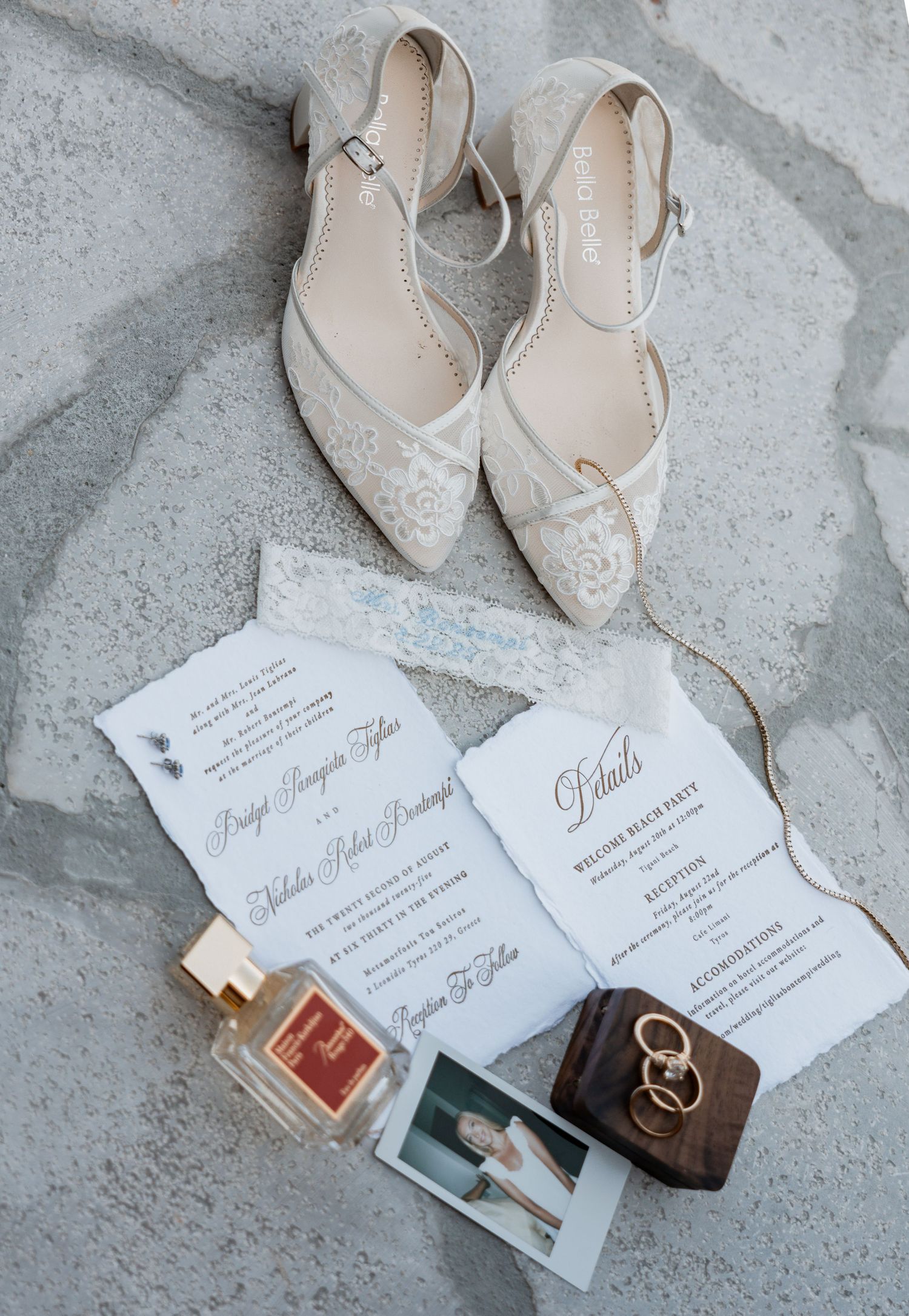 Elegant wedding flat shoes with lace detail beside invitation suite, perfume, rings, and polaroid photo on marble surface.