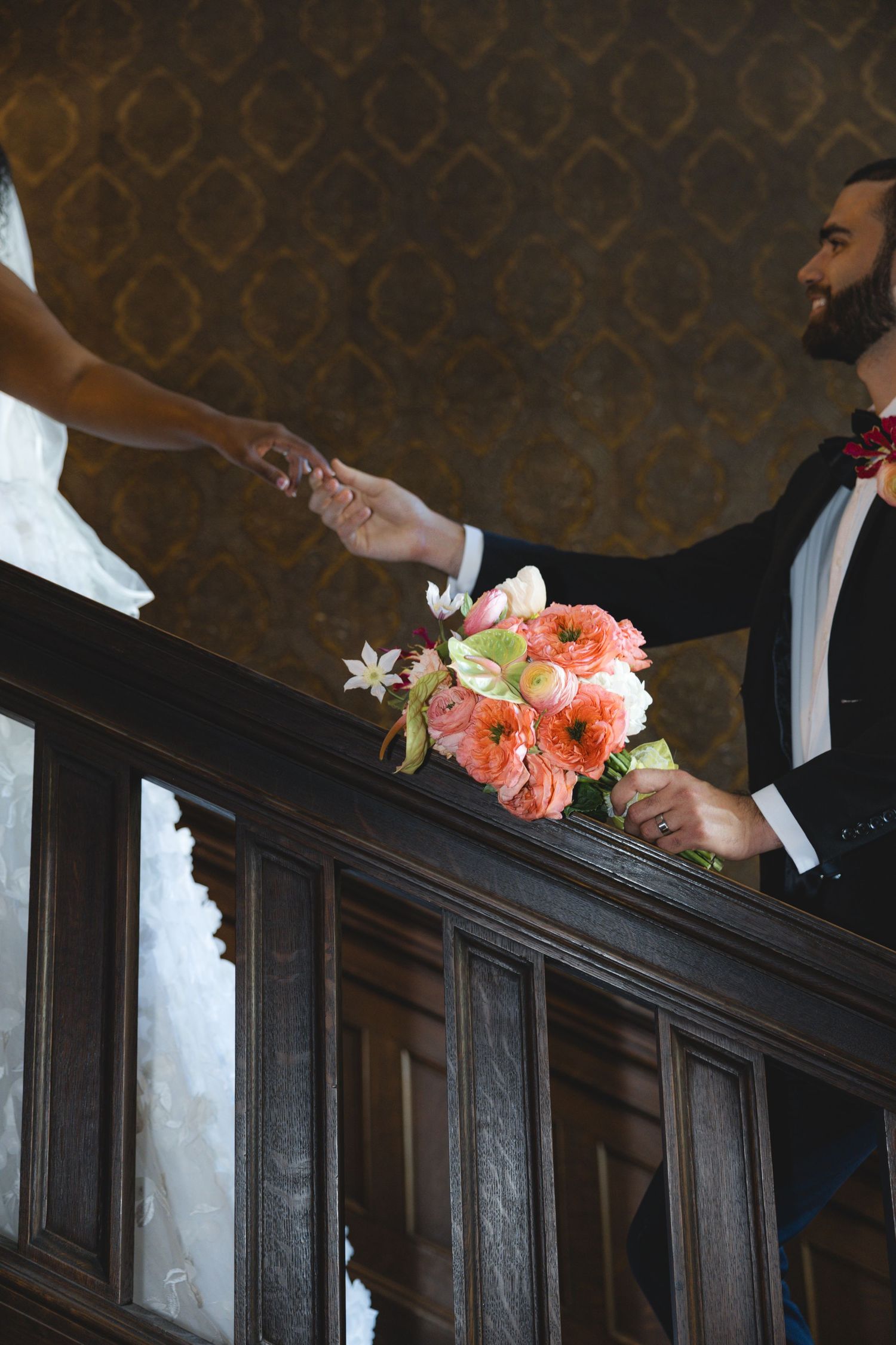 Groom holding bride's hand on ornate wooden staircase while carrying a coral and pink wedding bouquet.