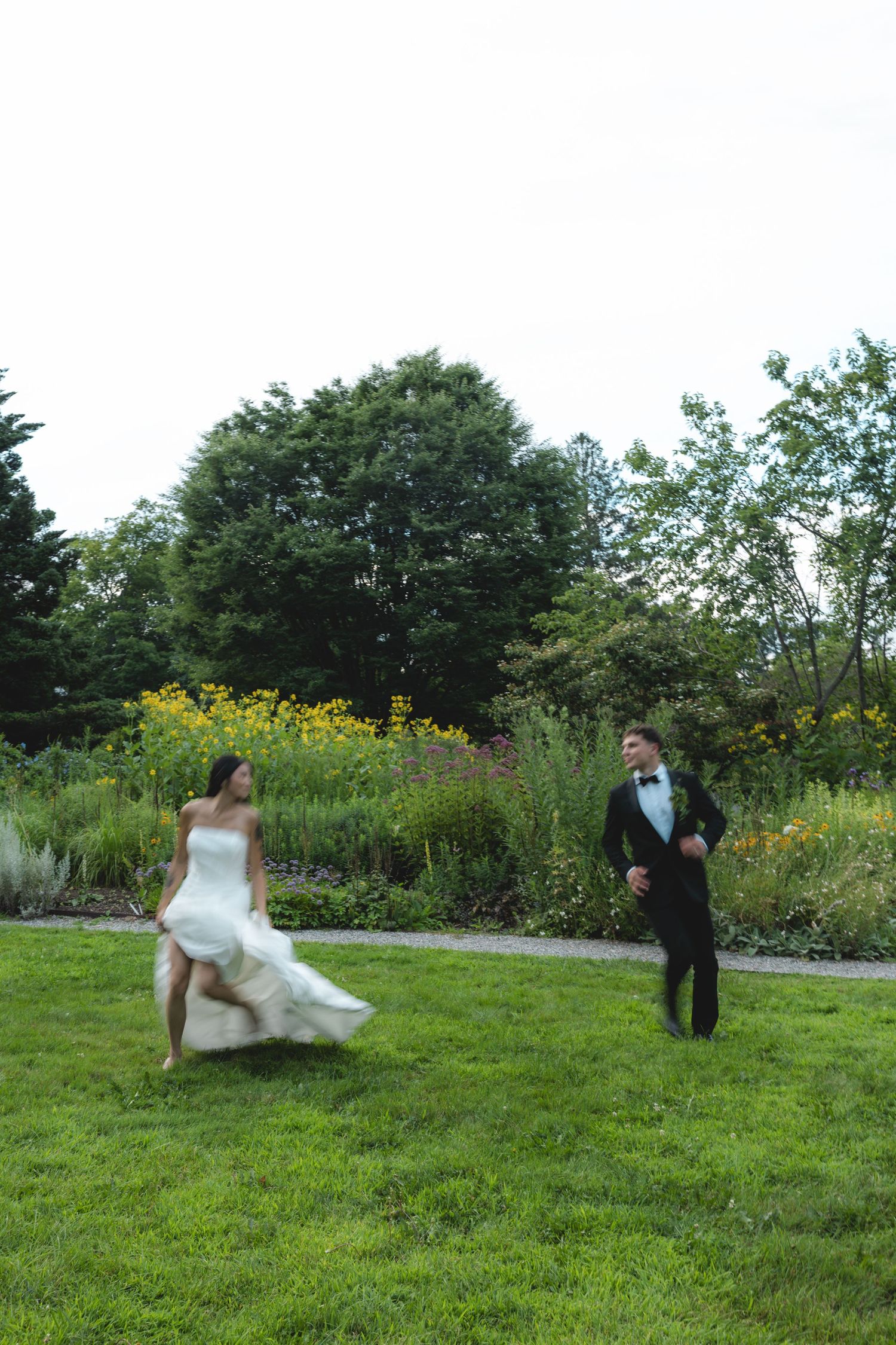Bride in white gown and groom in tuxedo running playfully on green lawn with colorful garden backdrop.