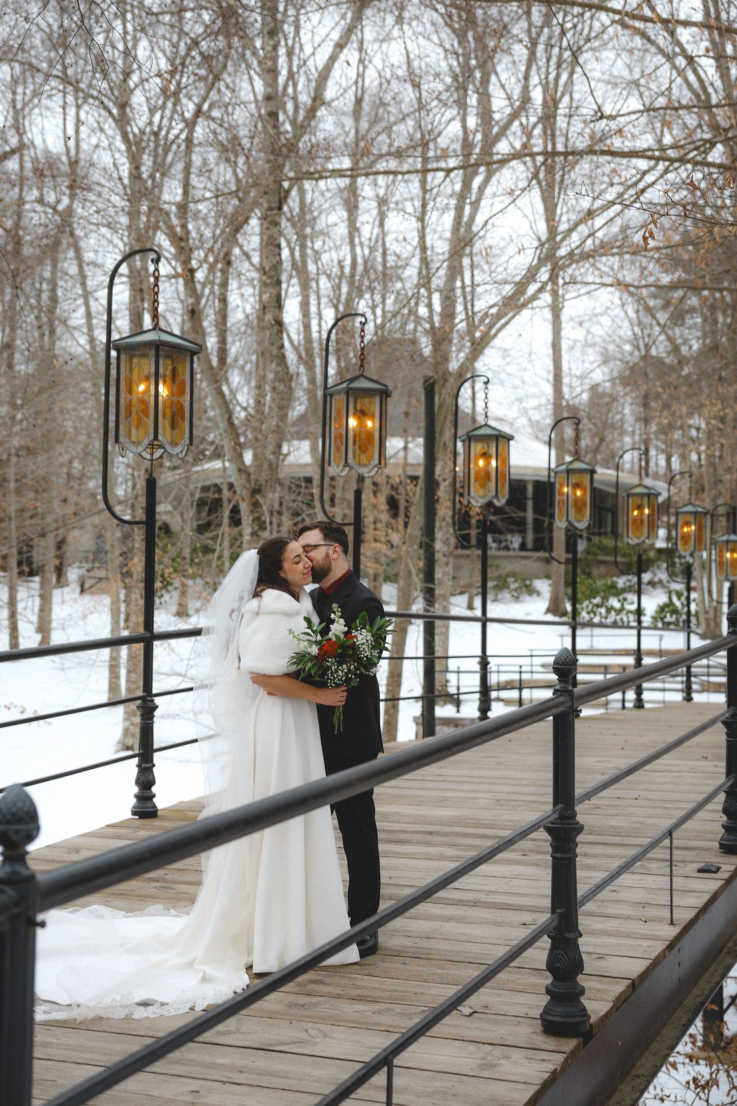 Winter wedding scene on a wooden bridge with glowing lanterns and snow-covered landscape.