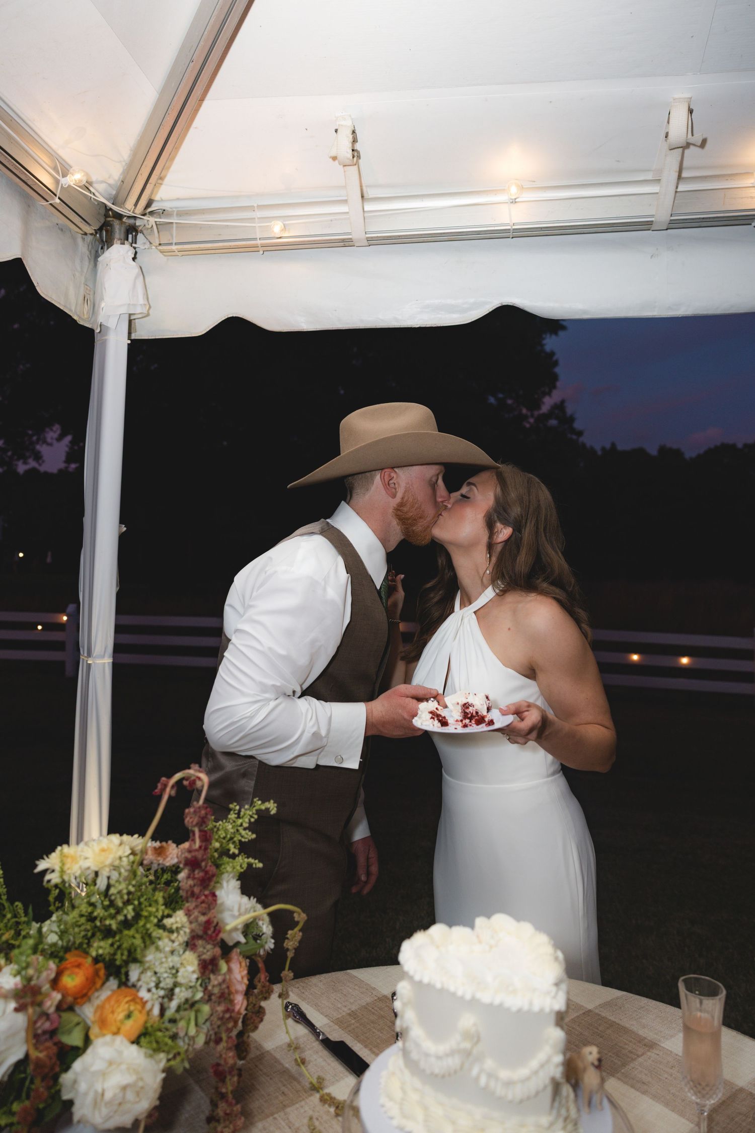 Bride and groom share a kiss while holding cake at their rustic outdoor wedding reception under a white tent.