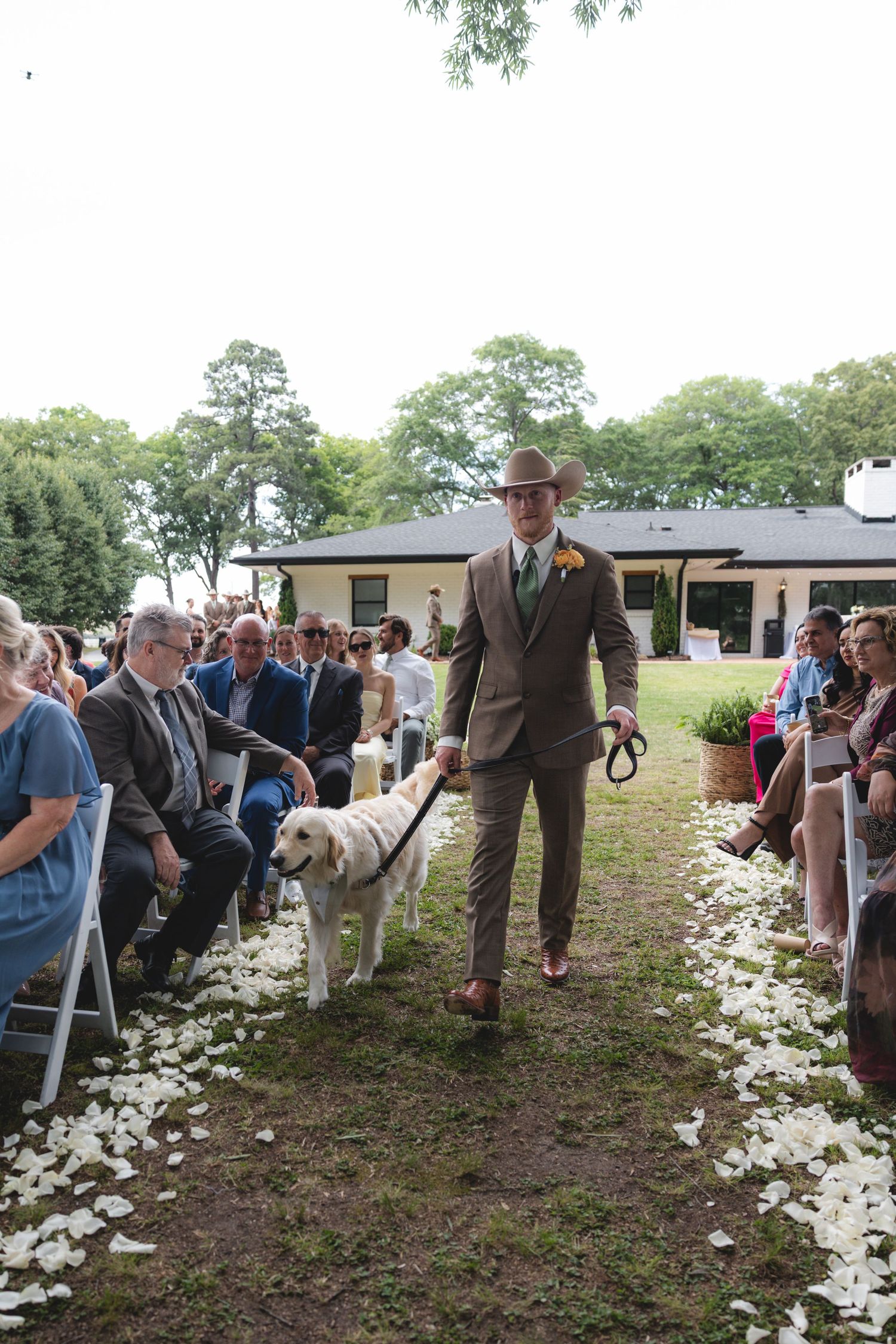 Groomsman in brown suit and cowboy hat walking yellow lab dog down white petal aisle.