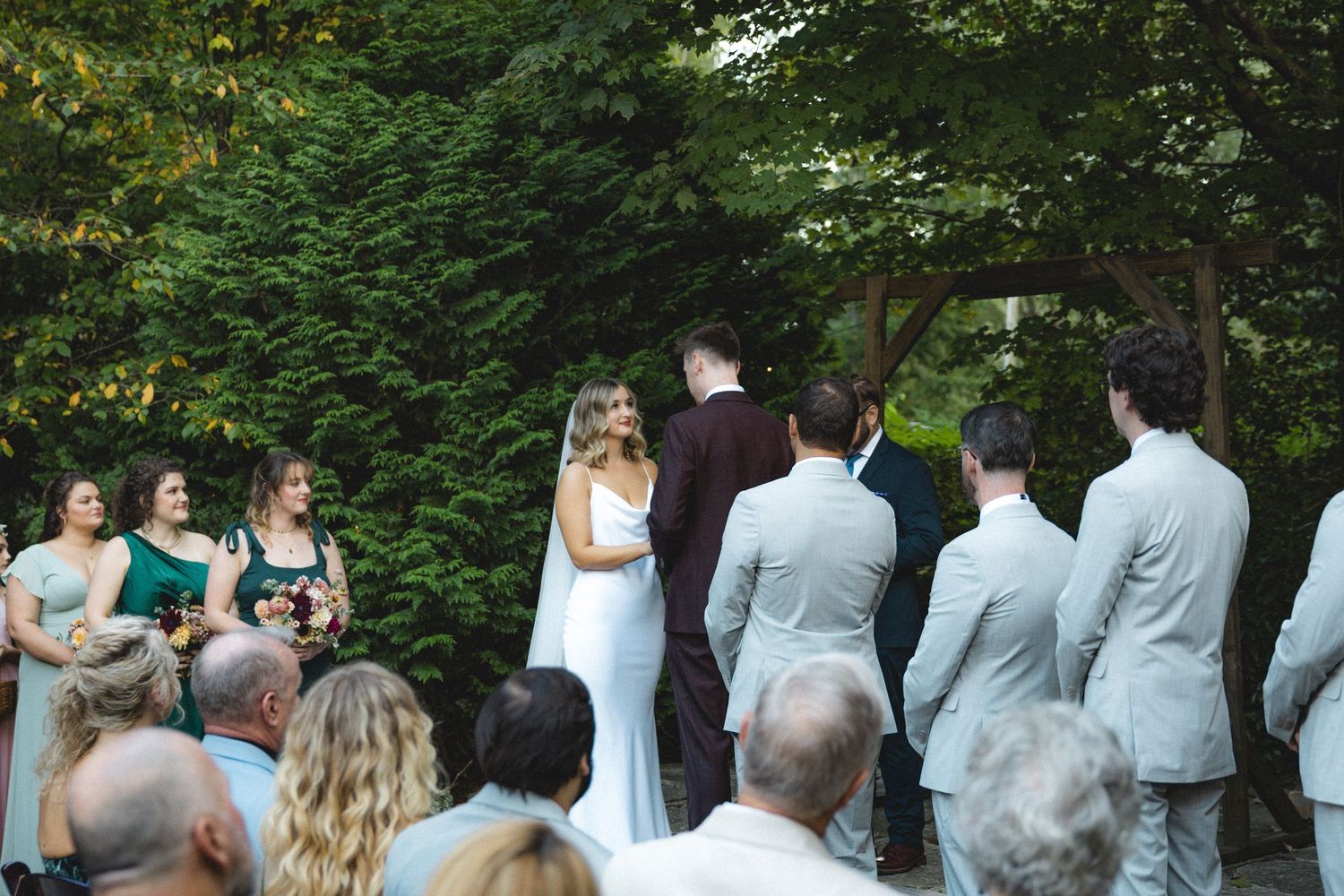 Outdoor wedding ceremony with bride and groom at the altar, surrounded by bridesmaids in green and groomsmen in gray suits.