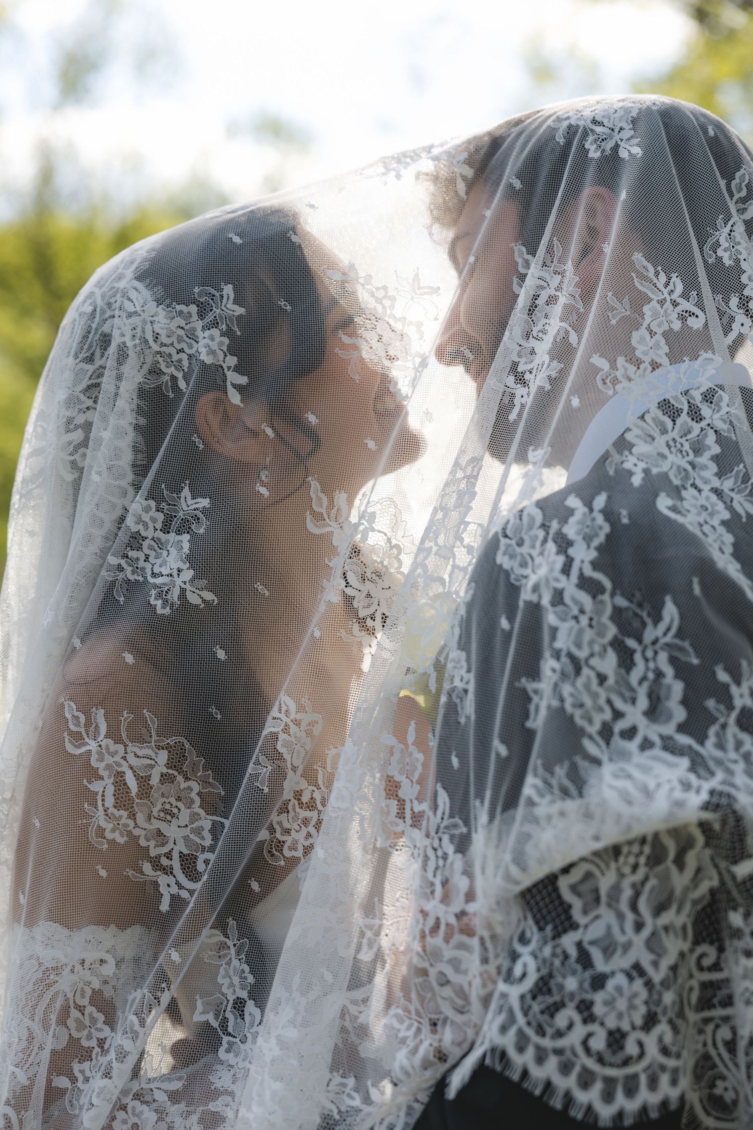 Bride and groom share intimate moment beneath lace bridal veil in romantic outdoor wedding photo.