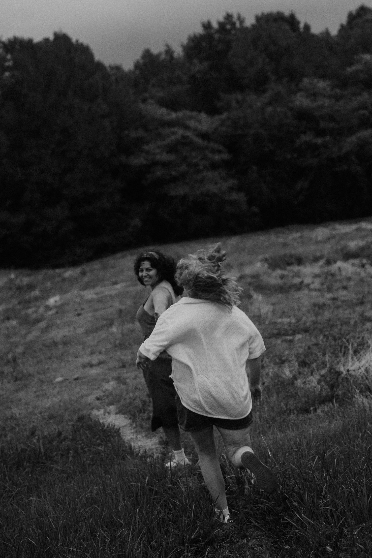 Two women running through a grassy hillside field in black and white photography with forest background.